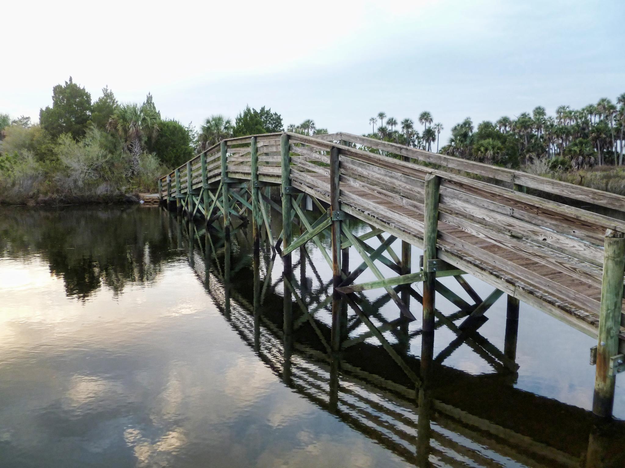 Long wooden bridge spanning calm water under an overcast sky, with trees.