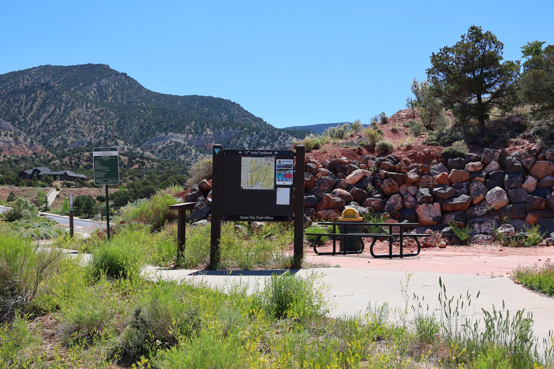 Information board and picnic table at a sunny, arid mountain trailhead.