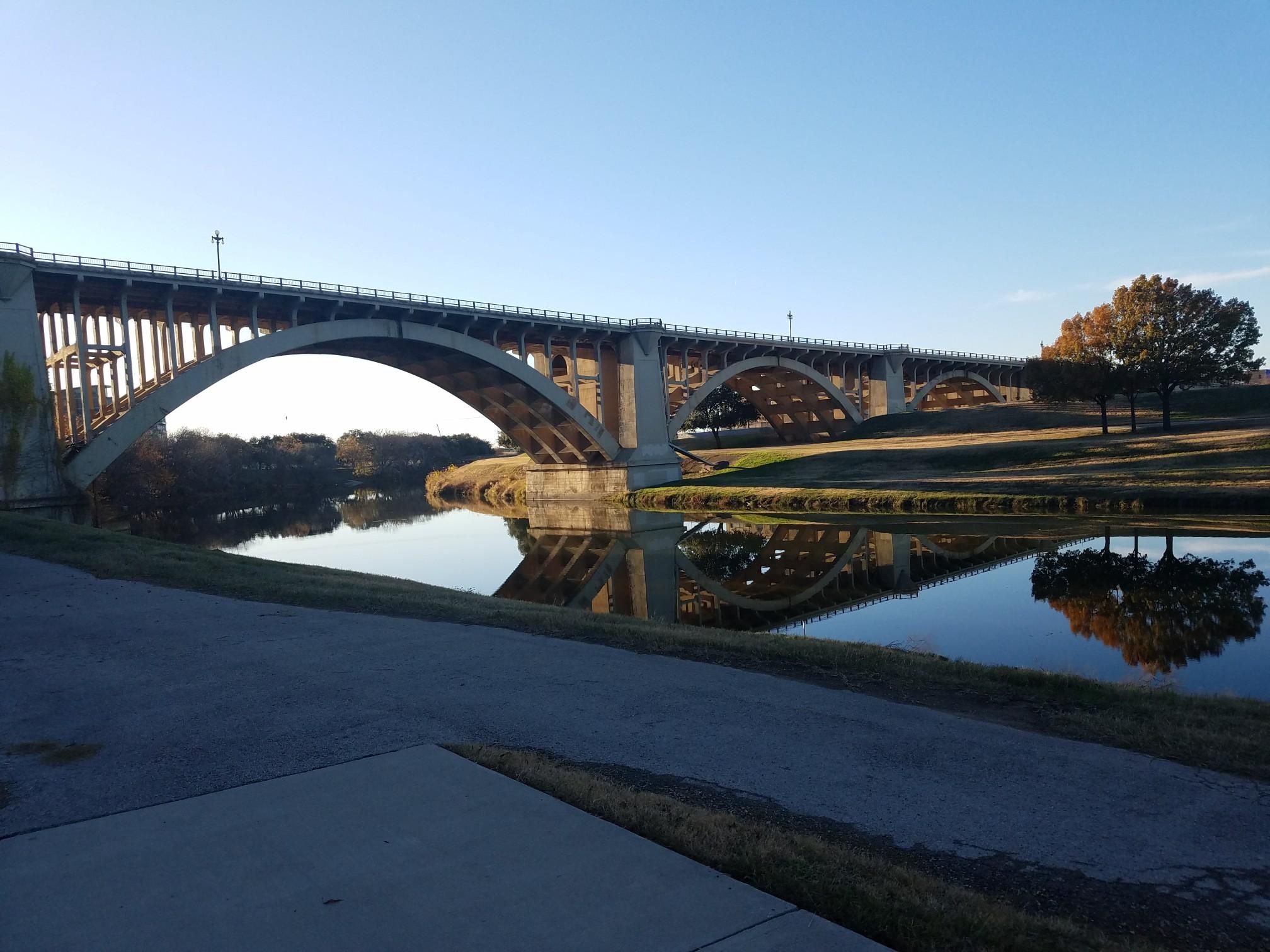 Arched concrete bridge over calm water reflecting the structure on a sunny day. - Trinity Trails: Stockyards and Downtown Loop
