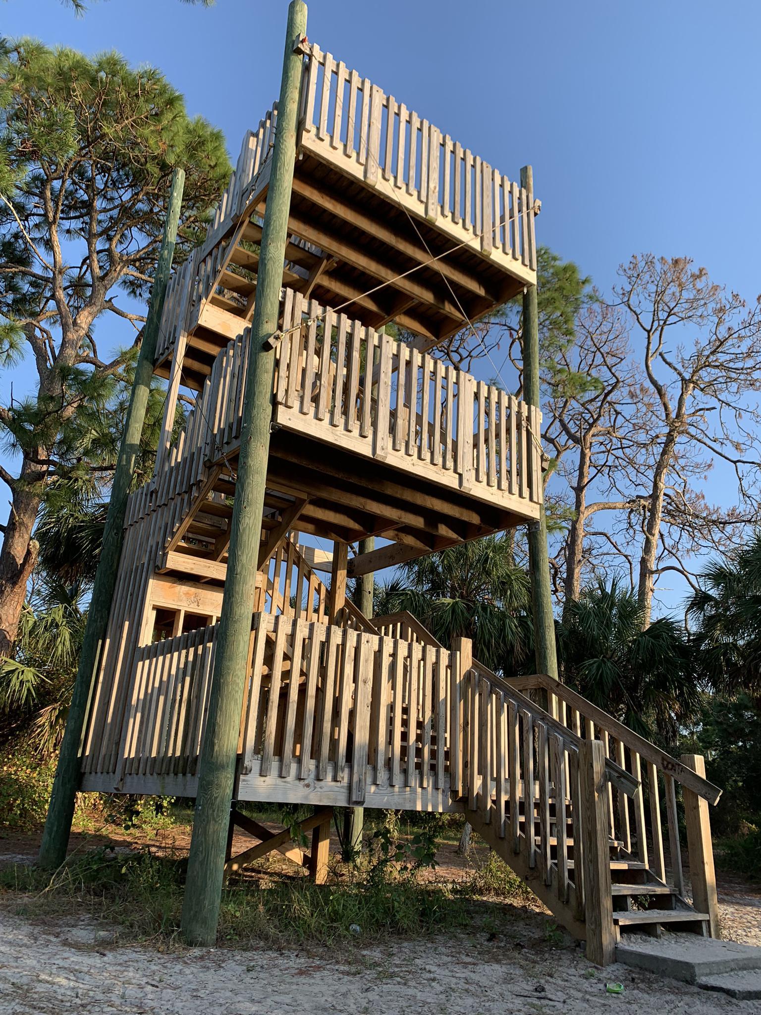 Tall wooden observation tower with multiple levels, surrounded by trees in bright light.