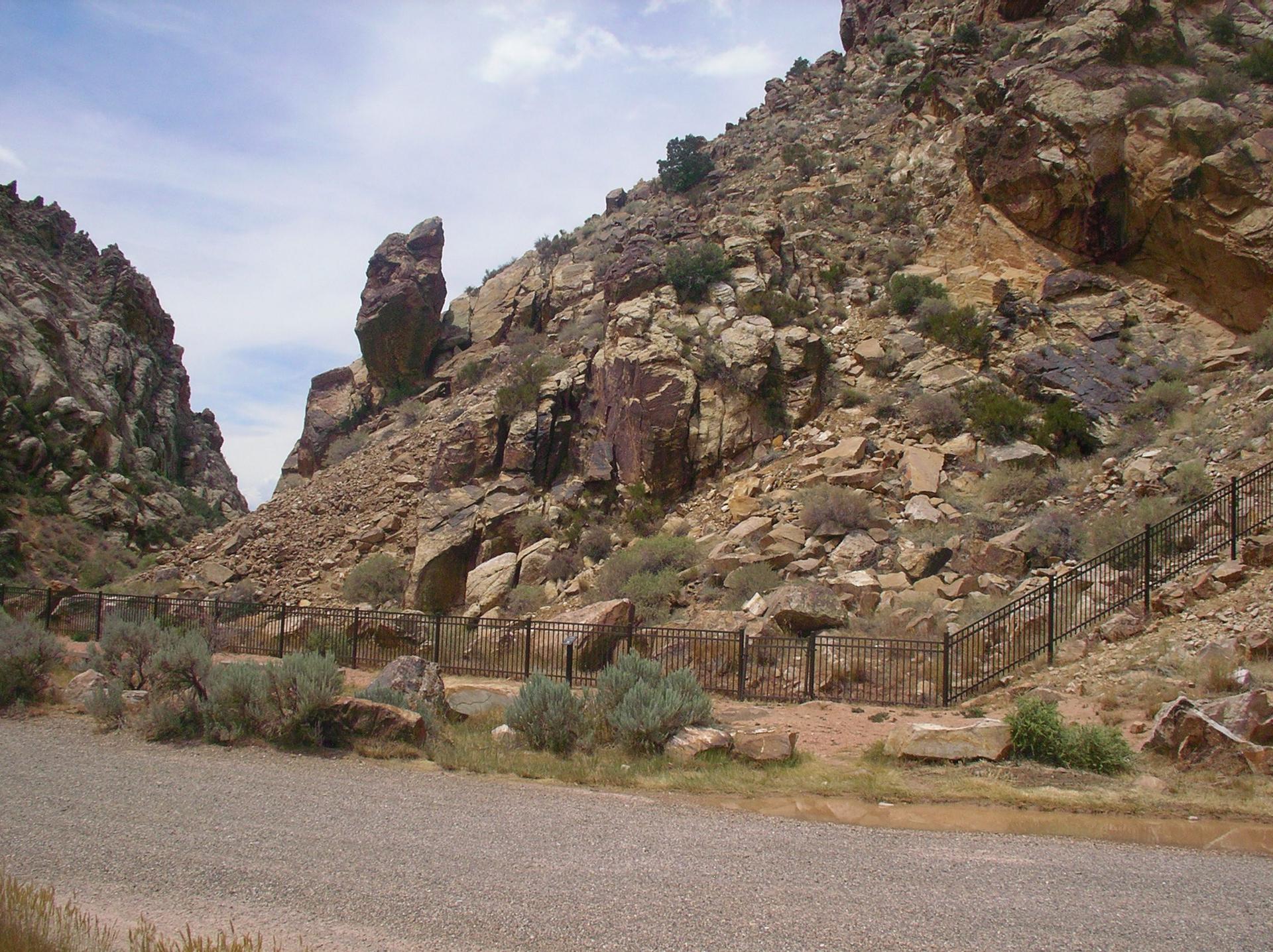 A gravel road runs alongside a metal fence at the base of a rocky canyon.