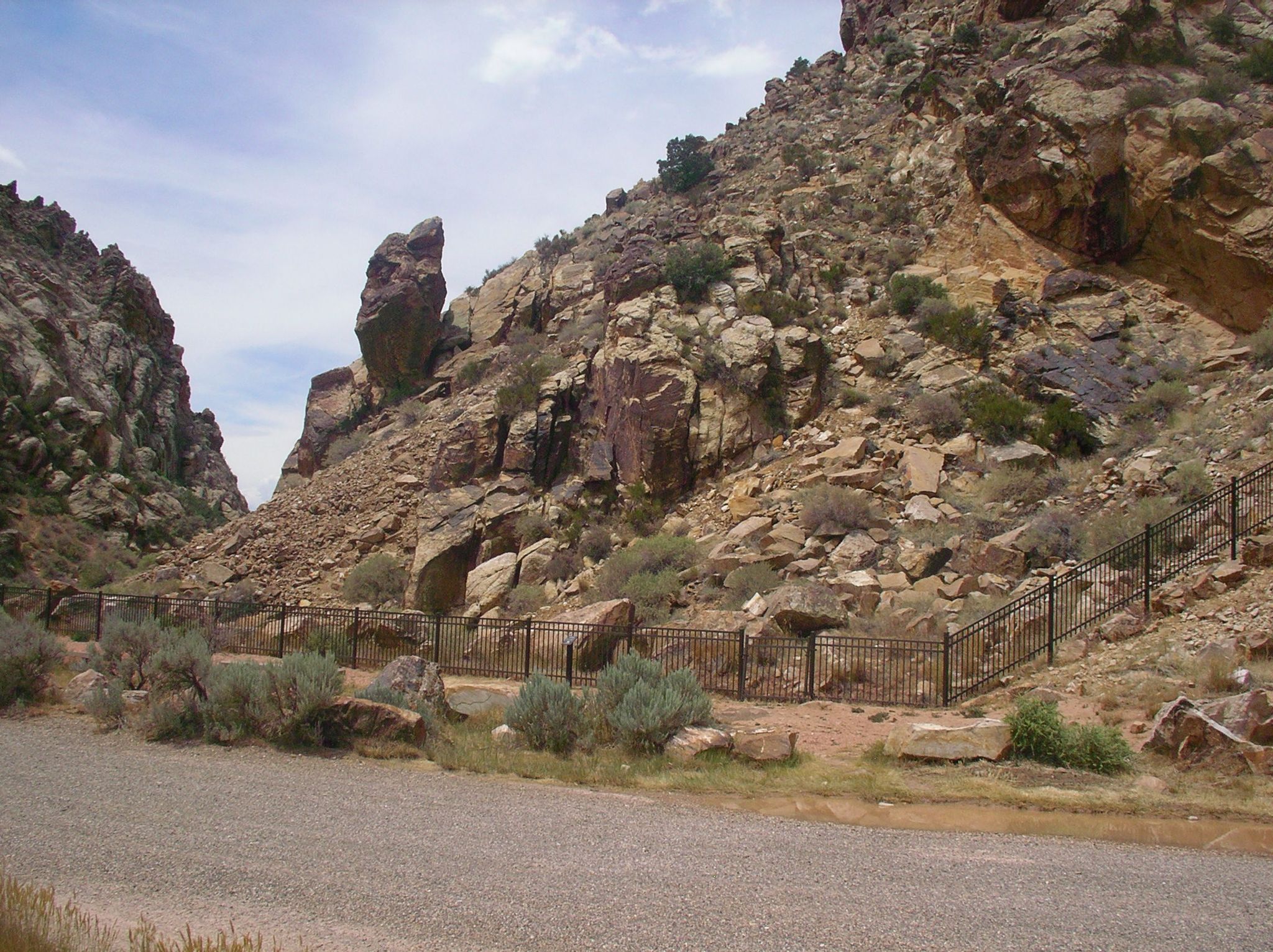A gravel road runs alongside a metal fence at the base of a rocky canyon. - Enoch to Parrowan loop