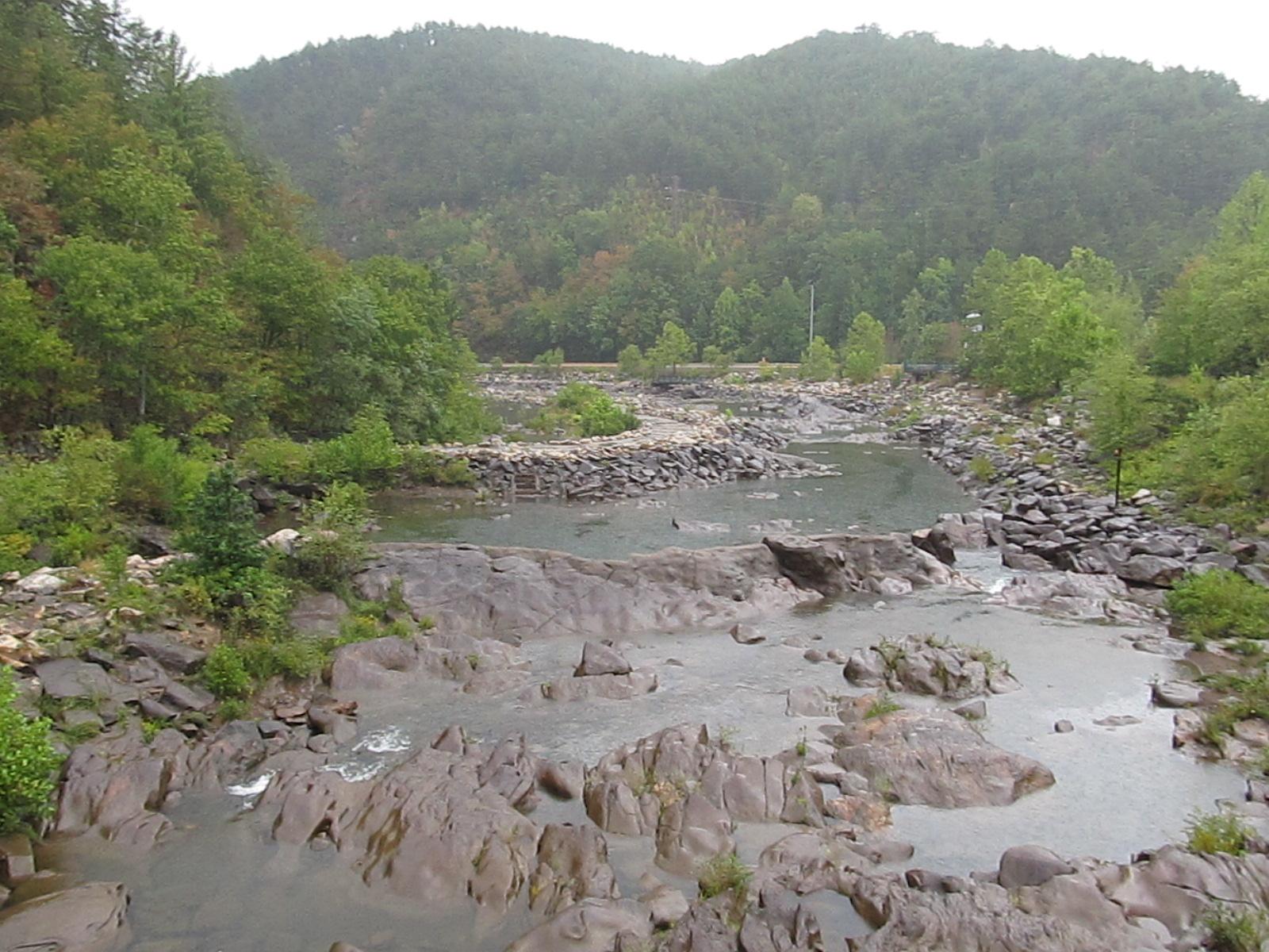 A wide, rocky river flows through a green, forested valley under an overcast sky.