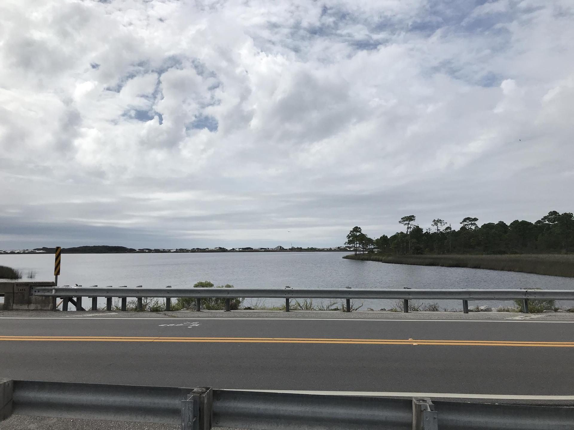 Roadside view of a wide bay with distant shoreline under a cloudy sky.