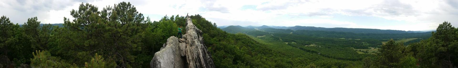 Panoramic view of a rocky ridge overlooking lush green mountains under cloudy skies.