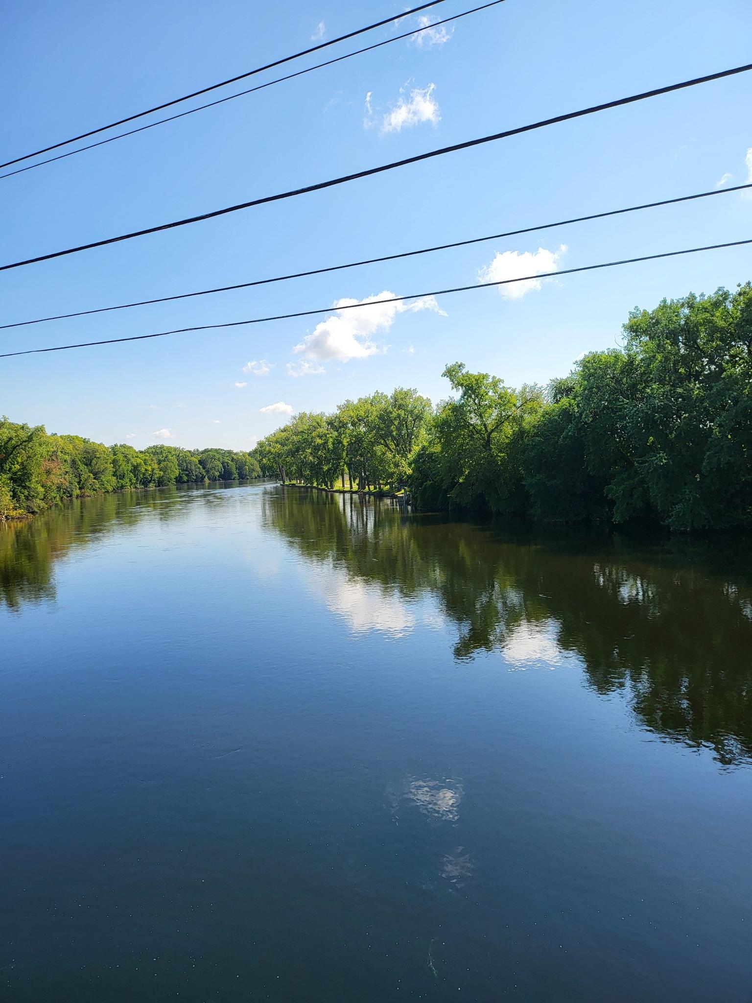 Calm river reflecting blue sky and green trees on a bright day.