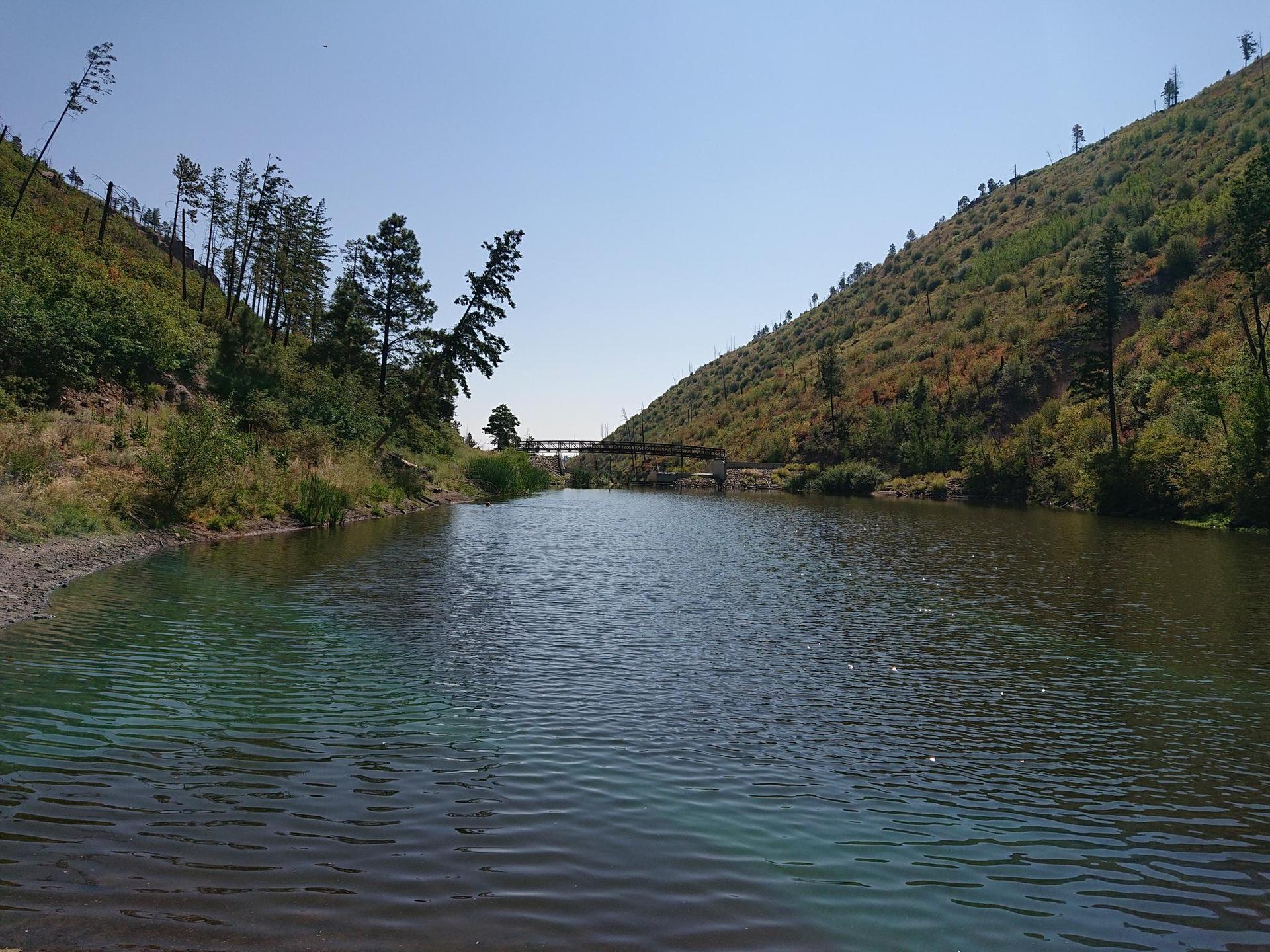 A calm river flows between green, tree-covered hills under a clear sky.