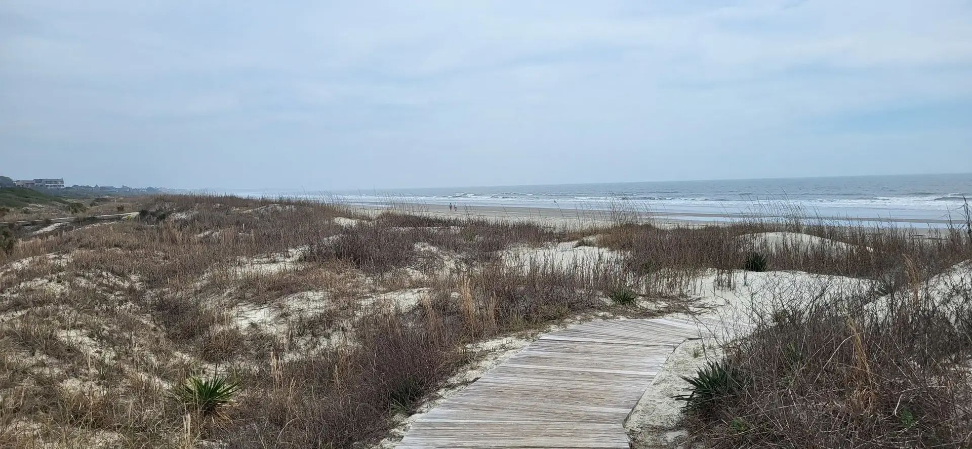 Wooden boardwalk through sandy dunes leads to a wide, overcast beach and ocean.