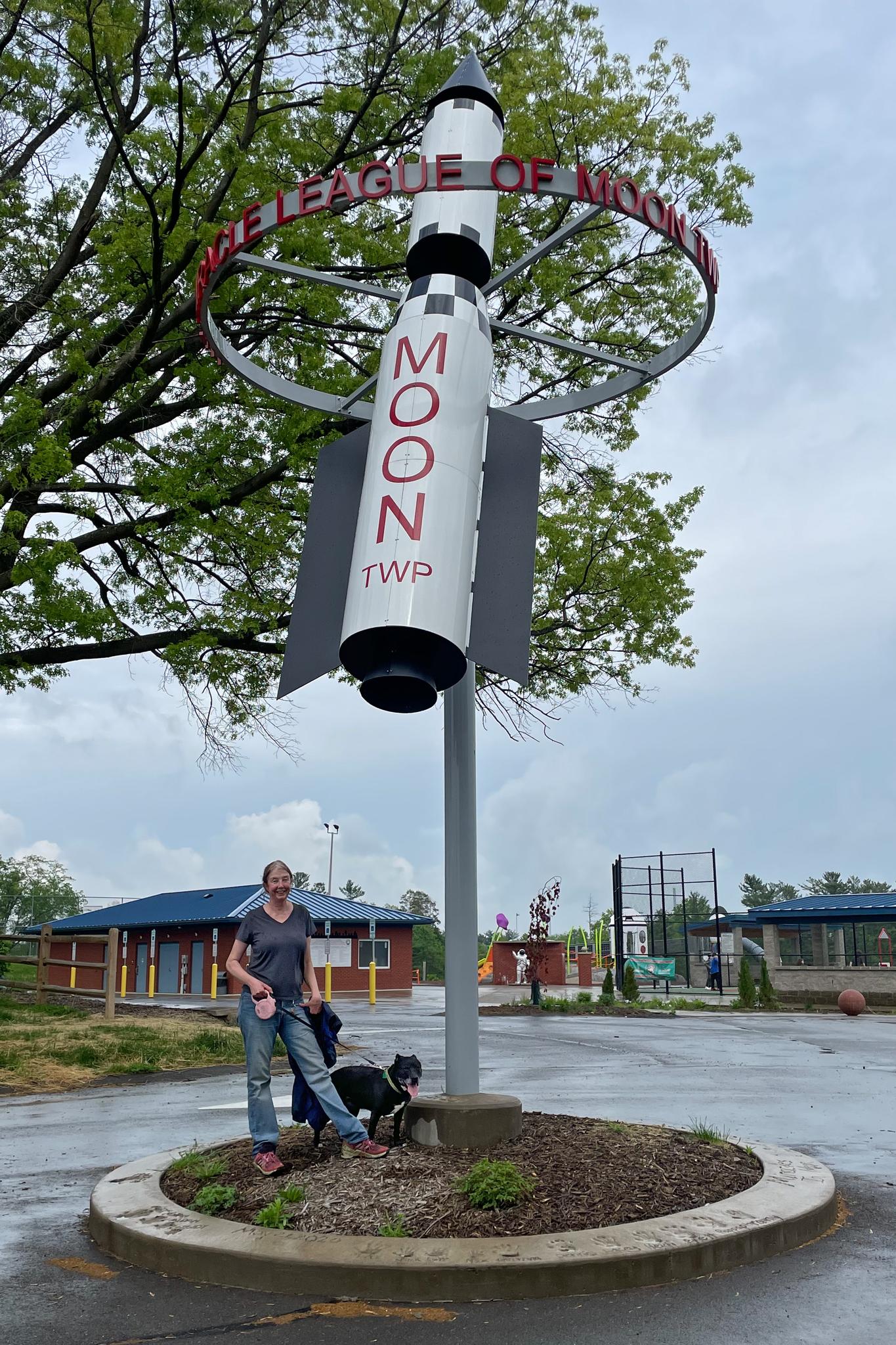 A person and dog stand by a large rocket sculpture on an overcast day.