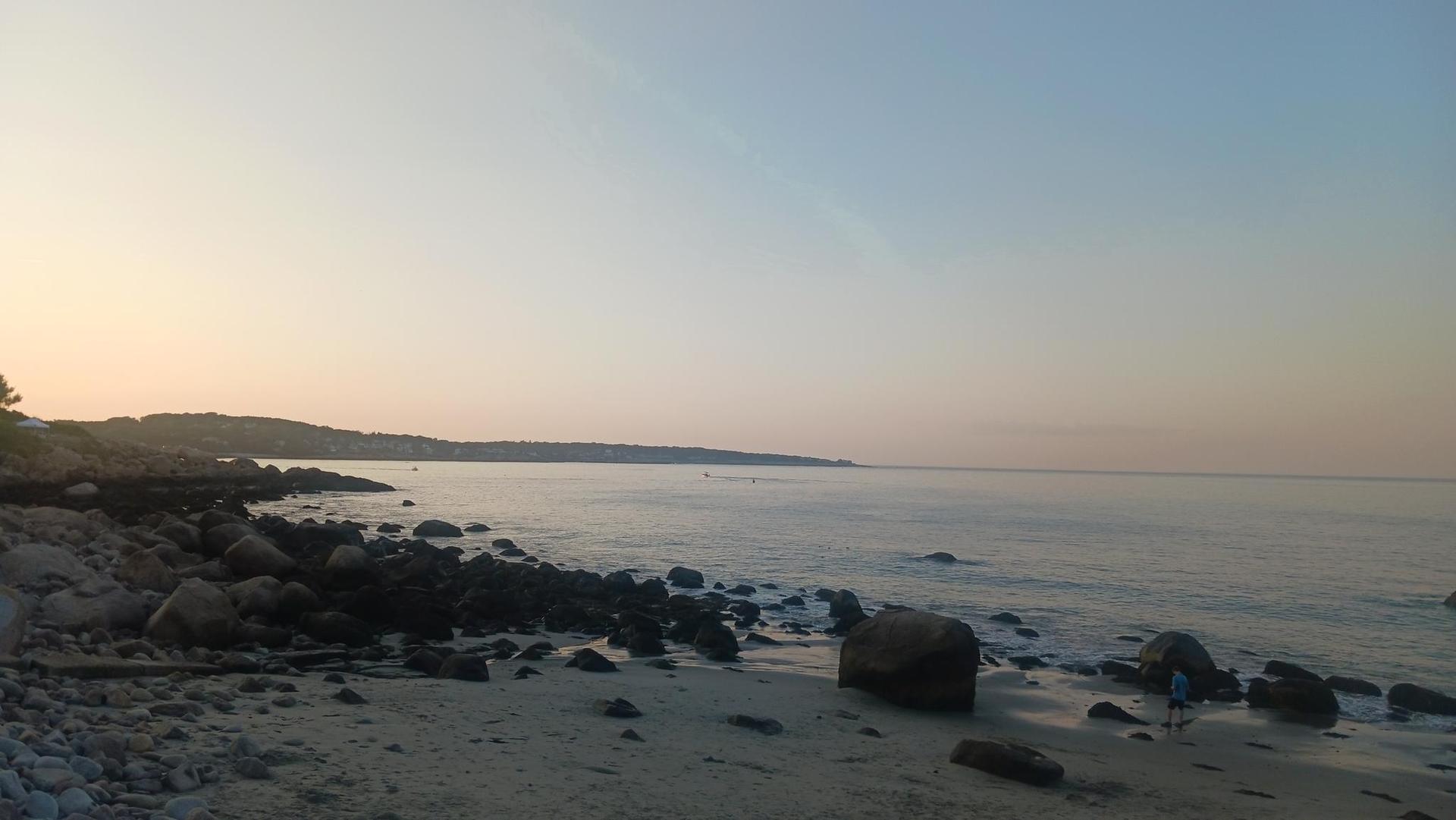 Calm ocean at sunrise with a rocky beach and distant land.