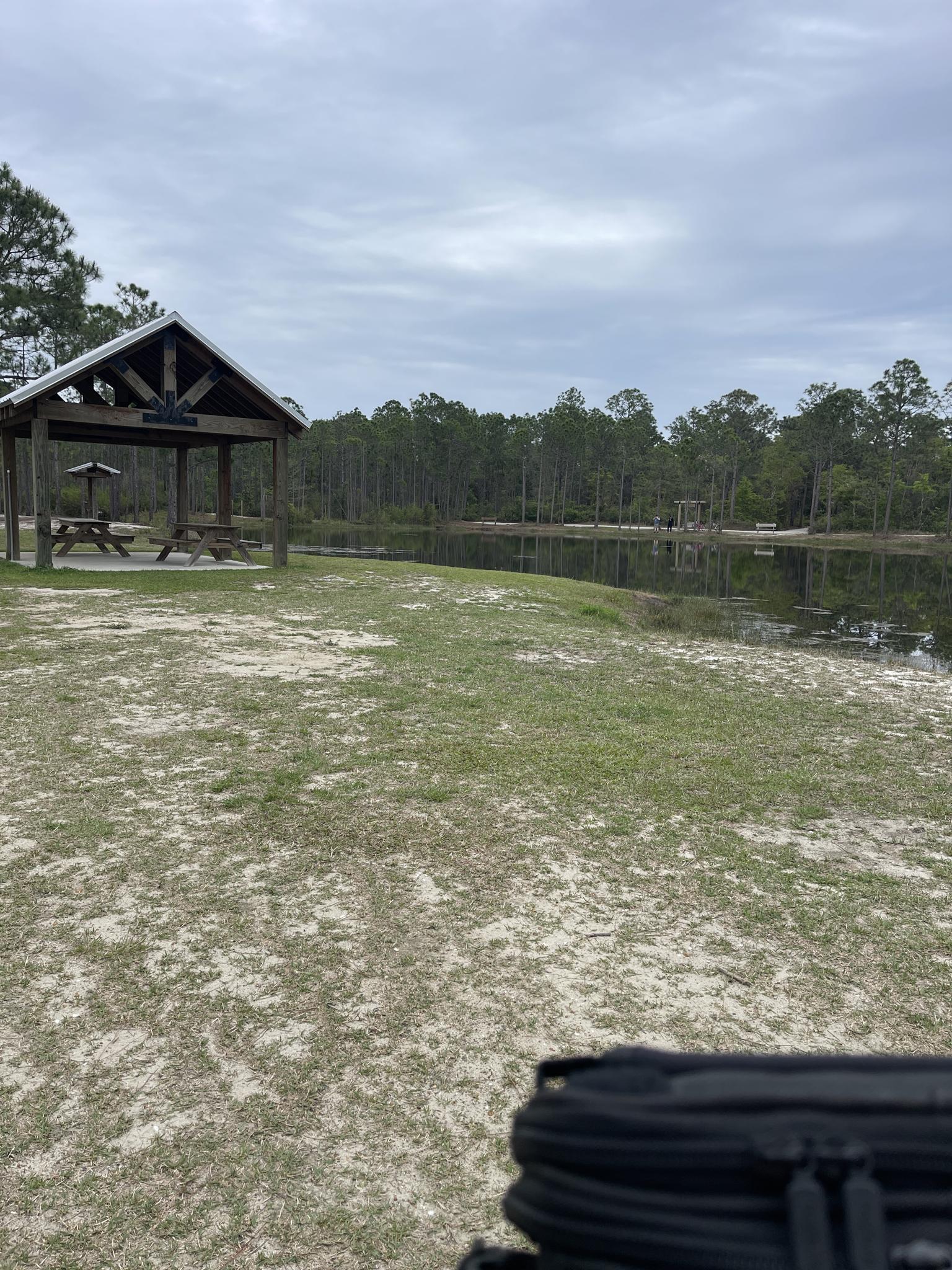 A wooden picnic pavilion stands by a calm lake under an overcast sky.