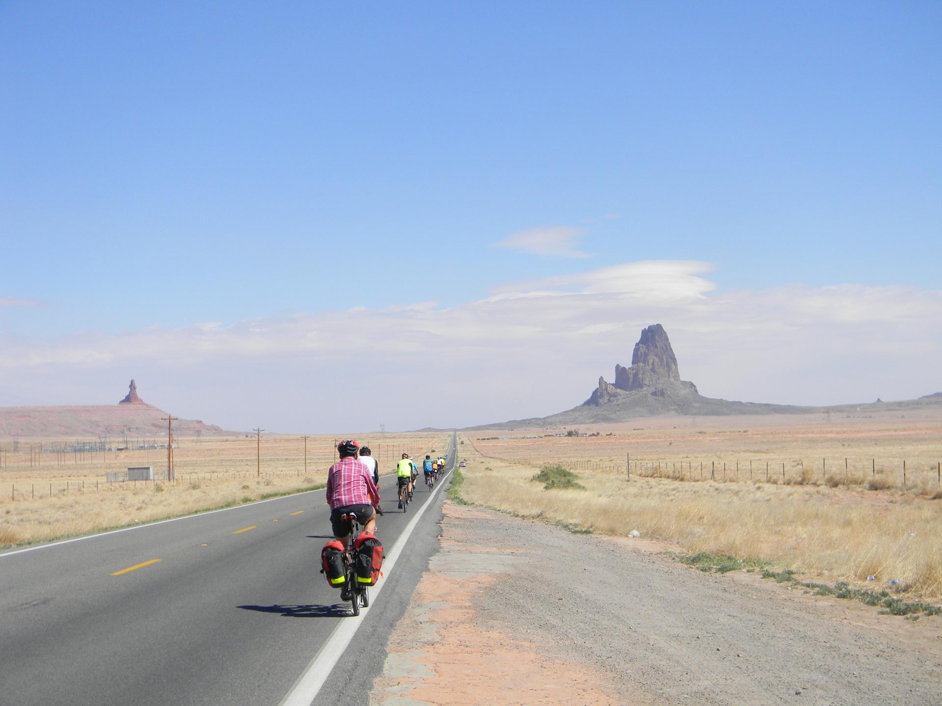 Cyclists ride on a paved road through a dry, flat landscape with distant peaks.