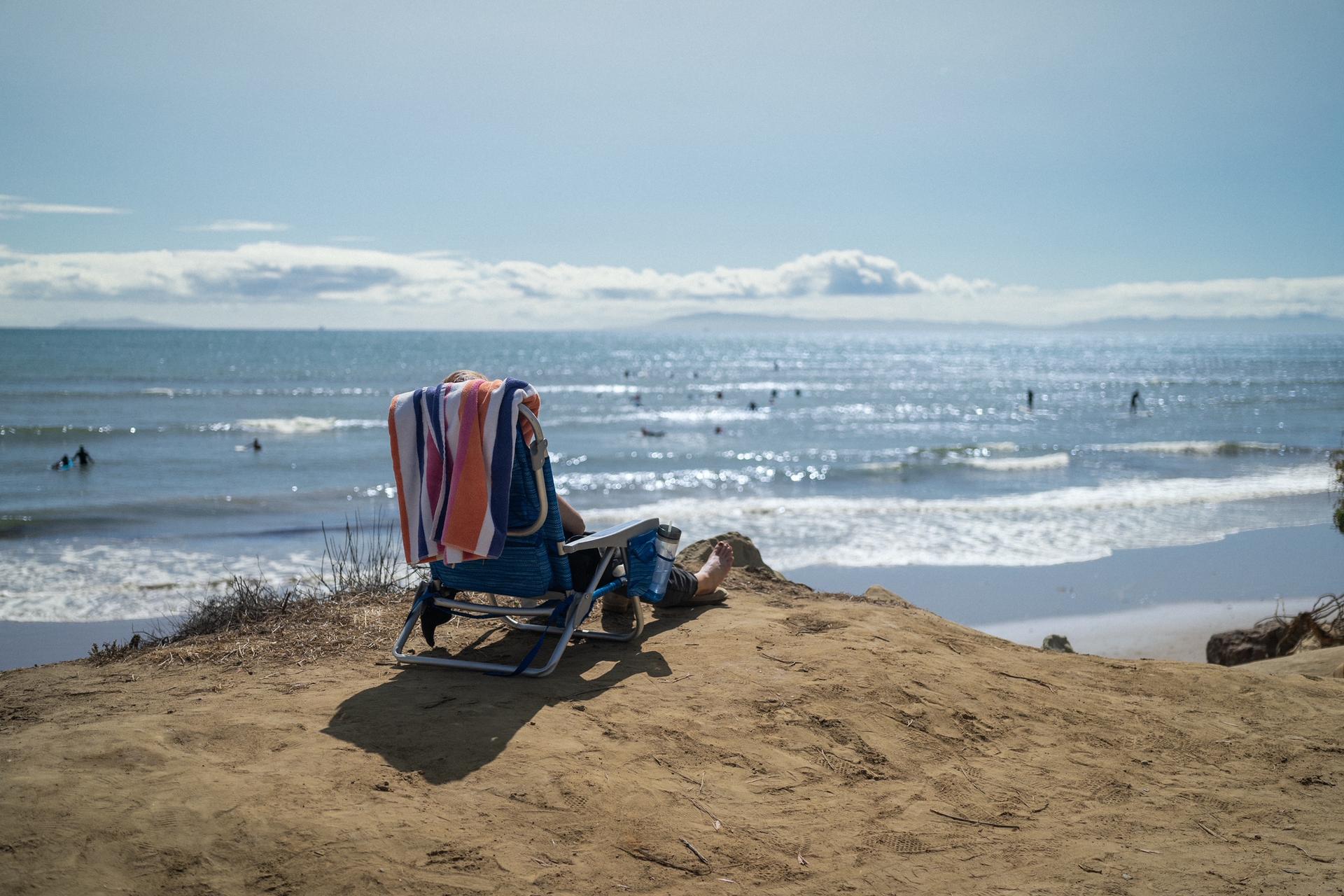 Person in beach chair on sandy bluff overlooking ocean with surfers, sunny day.