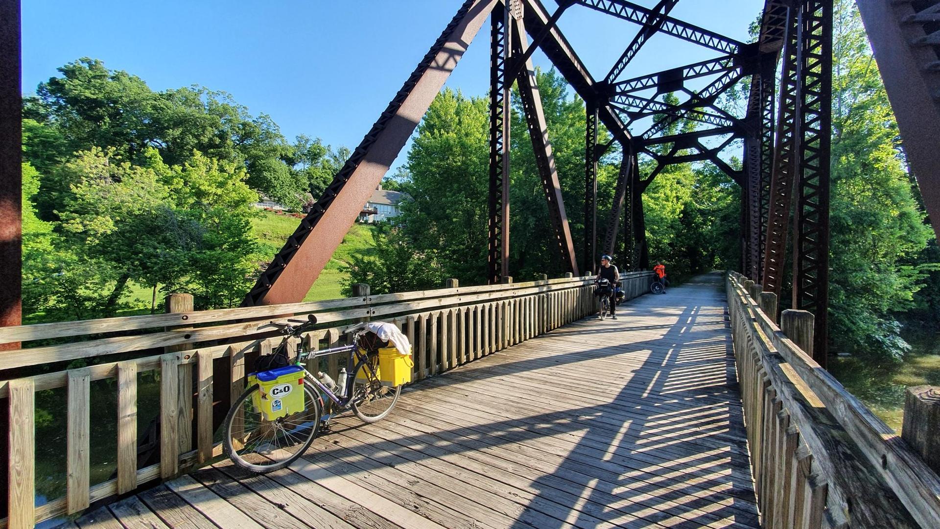 Cyclists on a wooden bridge with a brown metal truss, bright sunlight.