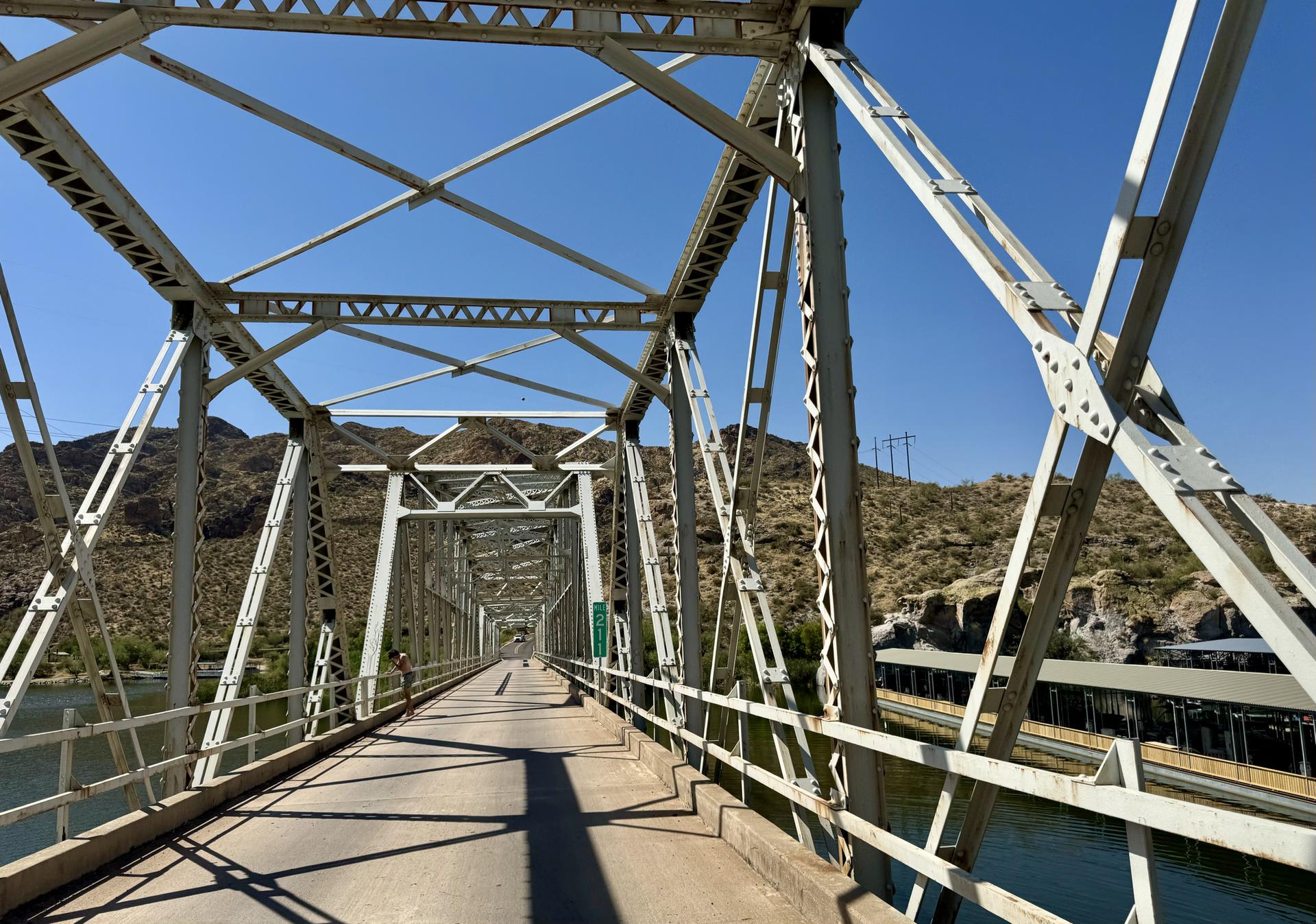 Metal truss bridge spanning over water with mountains in the background.