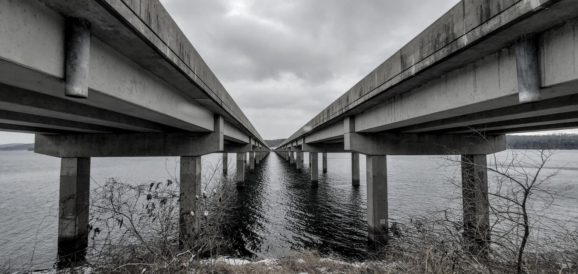 Underside of a long, concrete bridge stretching over dark water on an overcast day.