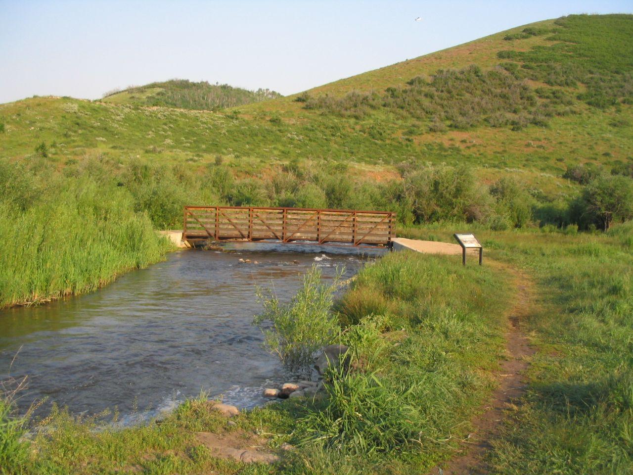 A wooden bridge spans a flowing stream amidst green hills on a sunny day.