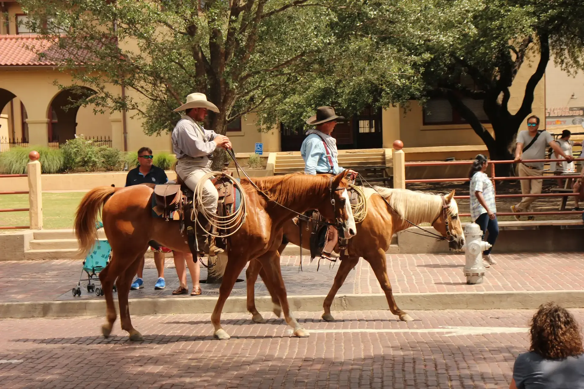 Two people on horseback ride down a brick street with spectators watching.