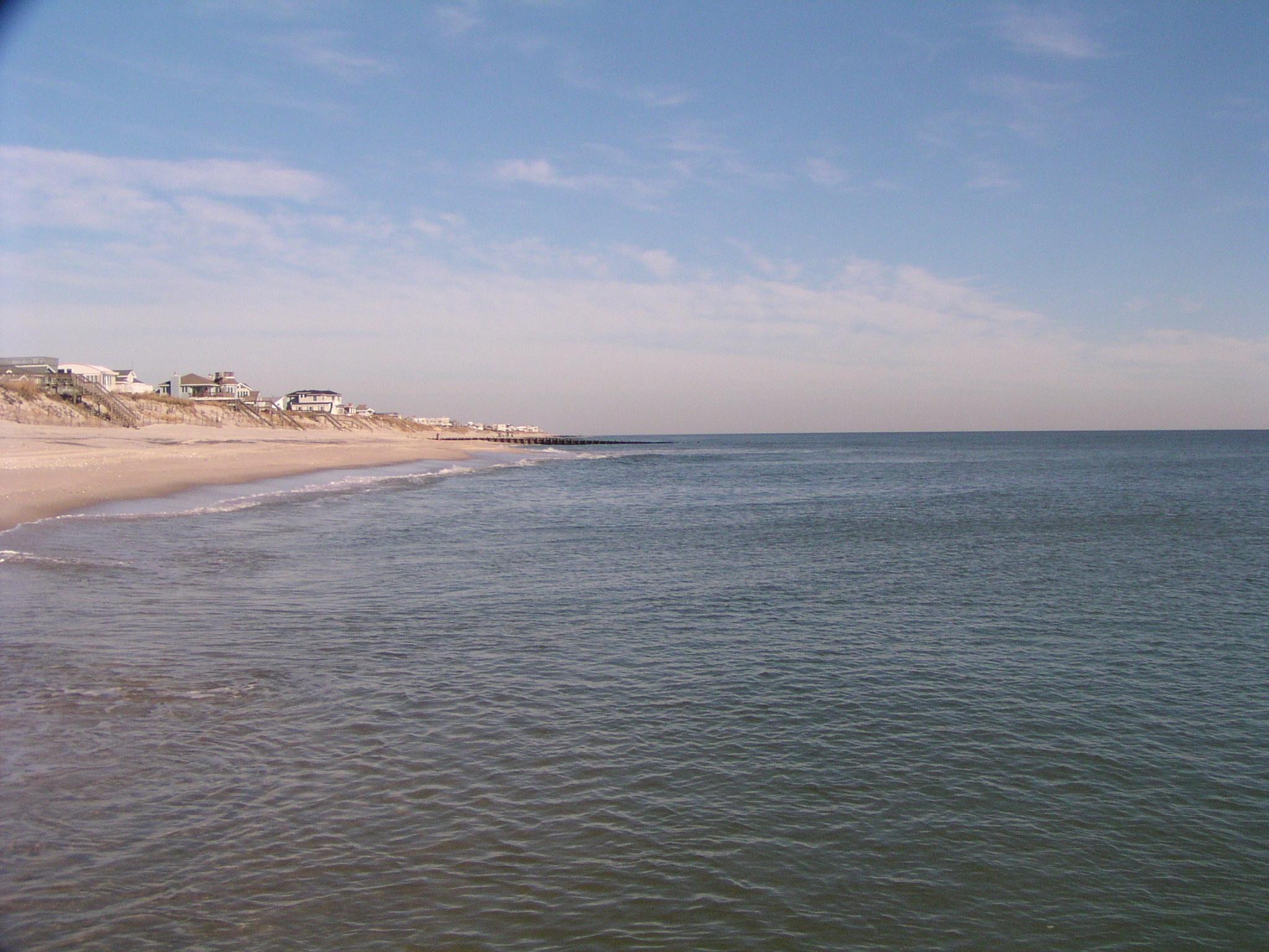 Calm ocean water meets a sandy beach with houses under a bright sky.