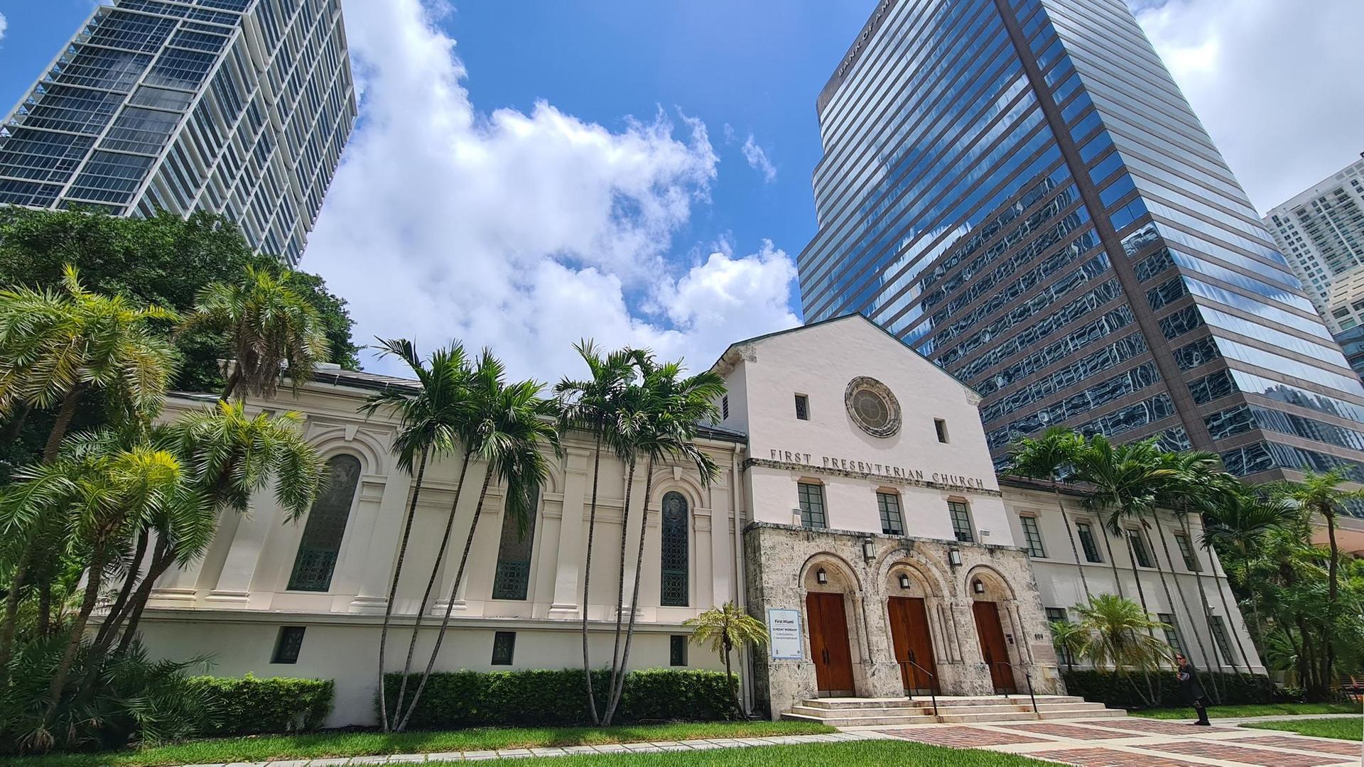 A historic building with palm trees, surrounded by modern skyscrapers on a sunny day.