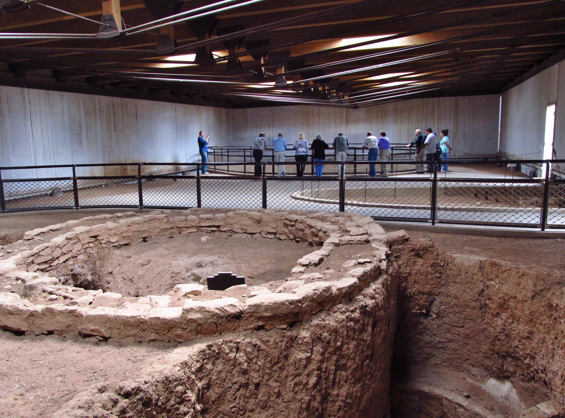 People observe excavated pit structures from a railing inside a building.