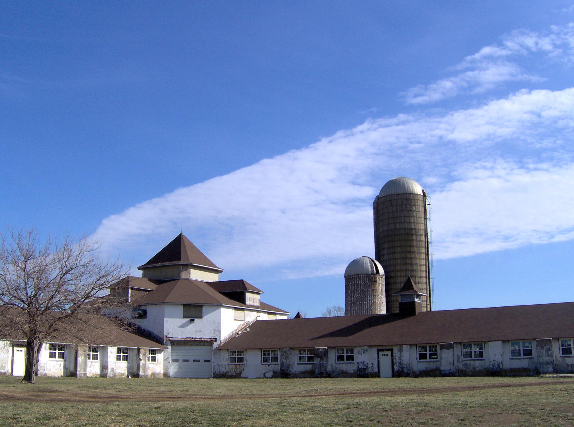 White farm buildings with two tall silos under a bright, cloudy blue sky.