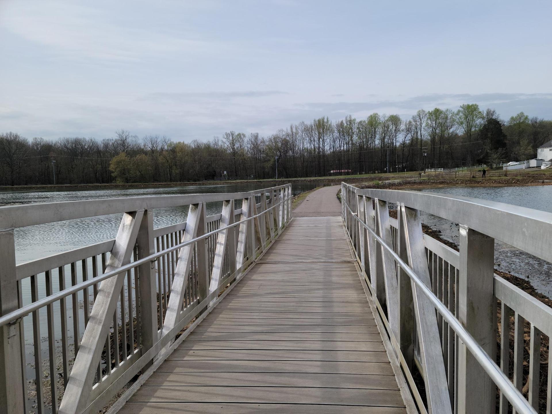A wooden and metal bridge spans calm water under a cloudy sky.