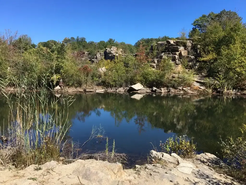 A calm lake reflecting trees and rocks under a clear blue sky.