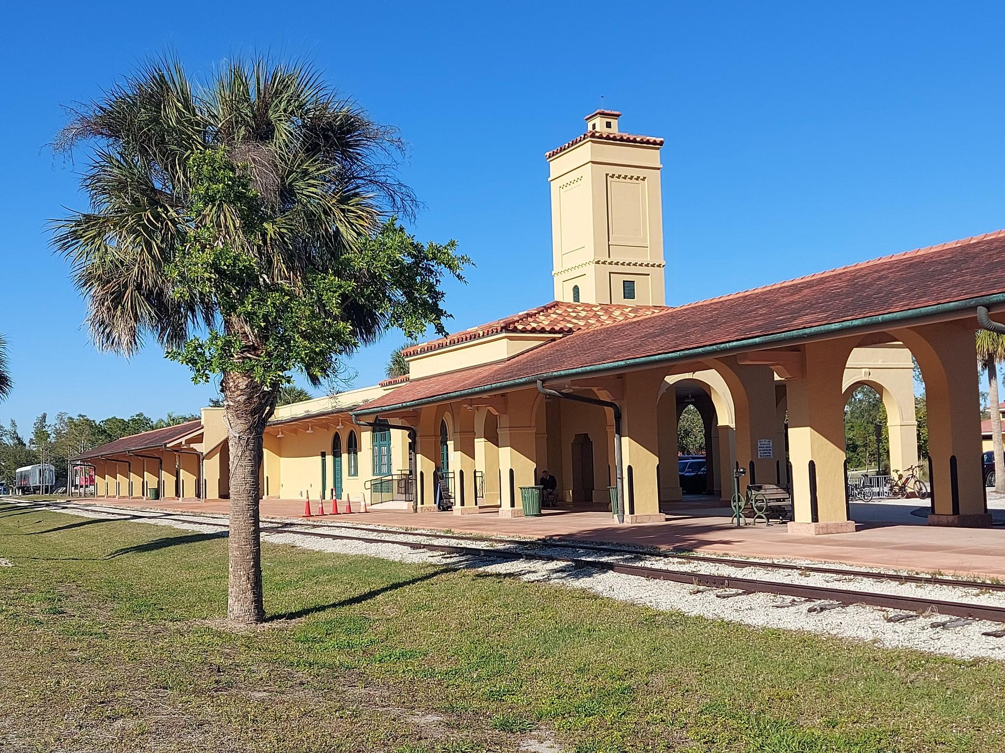 Old-style building with a tower next to railroad tracks and a palm tree.