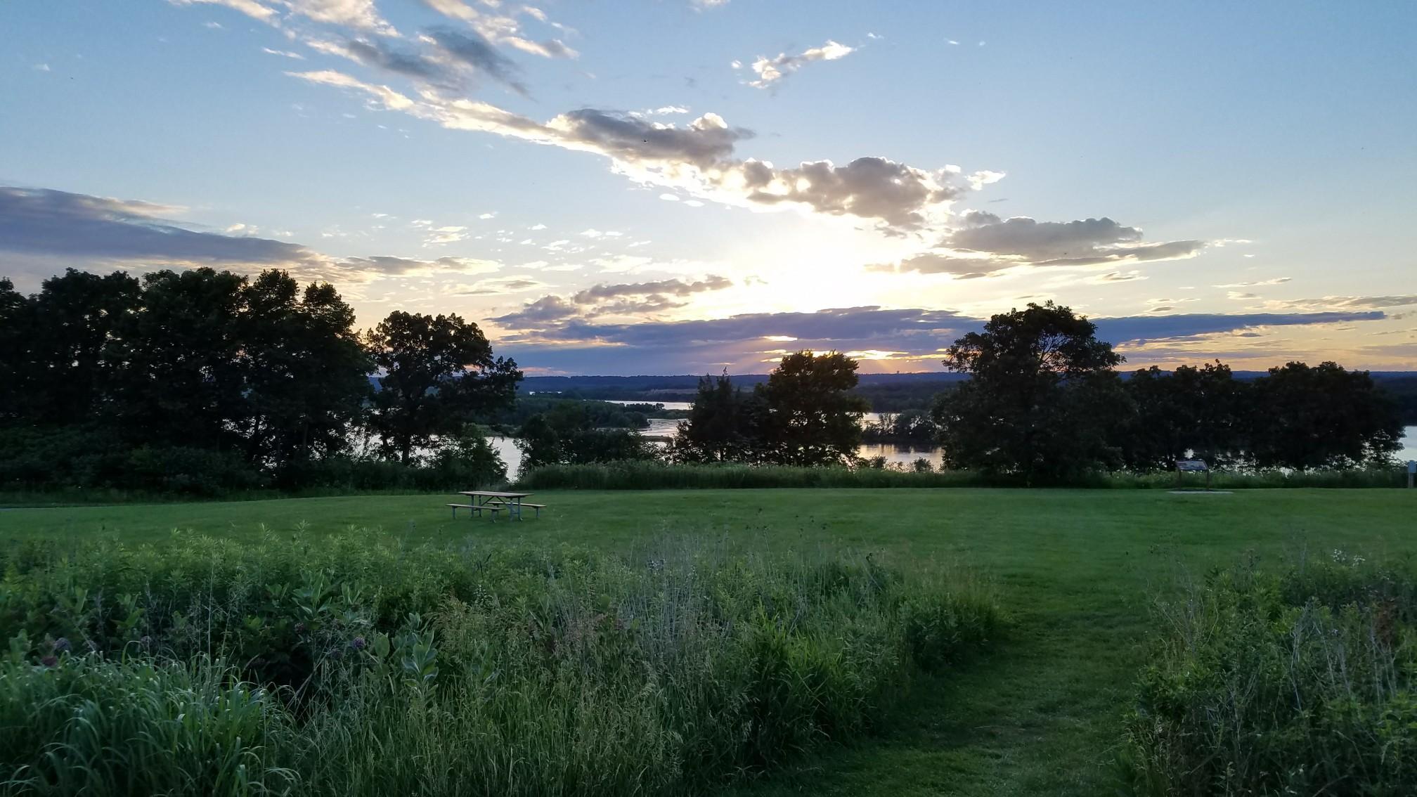 Grassy field overlooking a river at sunset, with a picnic table.