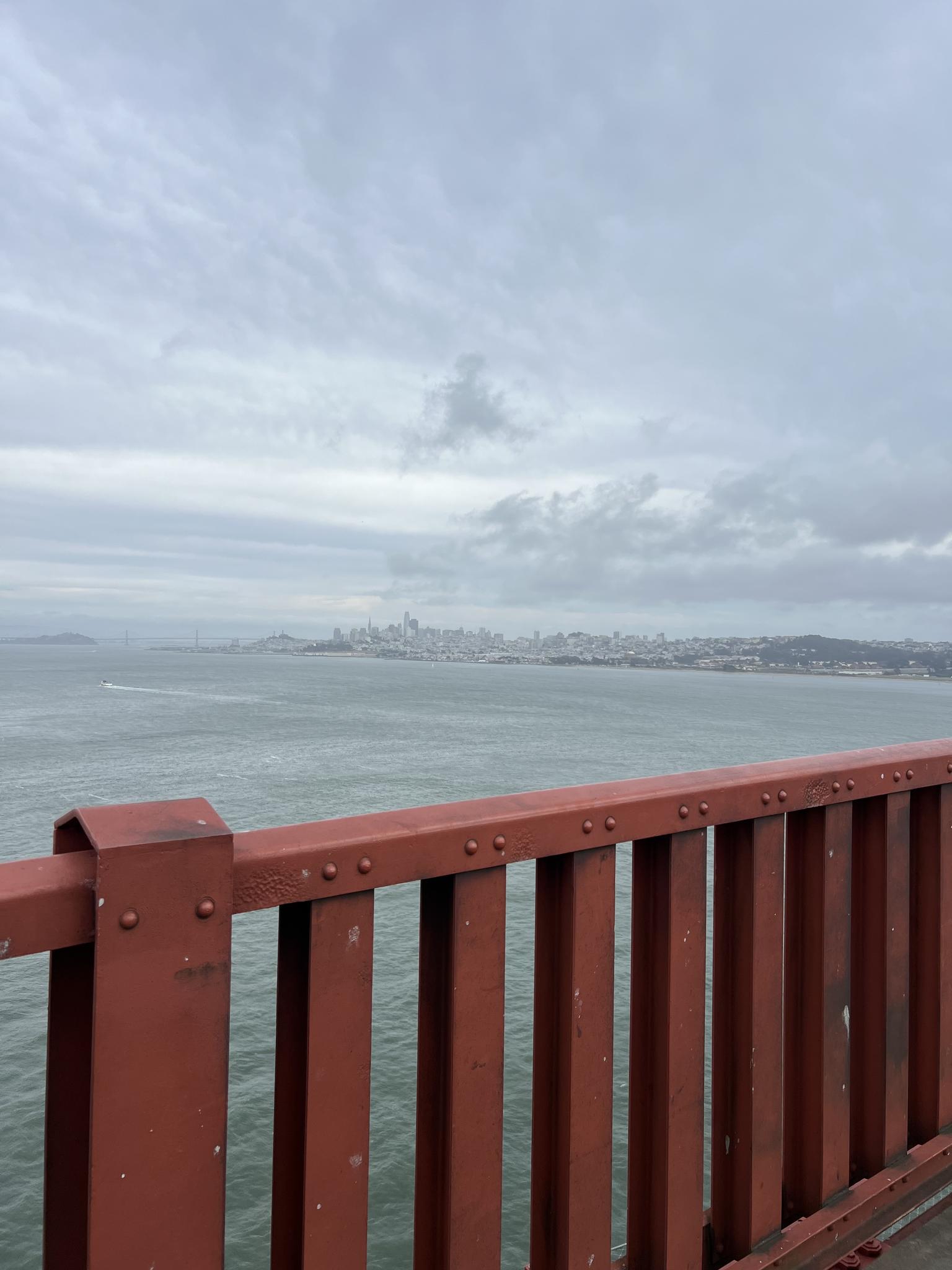 Red bridge railing, city skyline across water under cloudy sky.