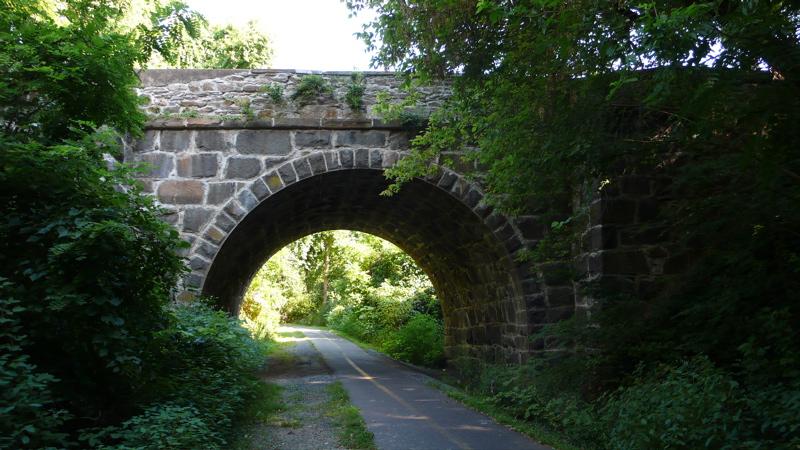 A stone arch bridge over a paved path surrounded by green trees.