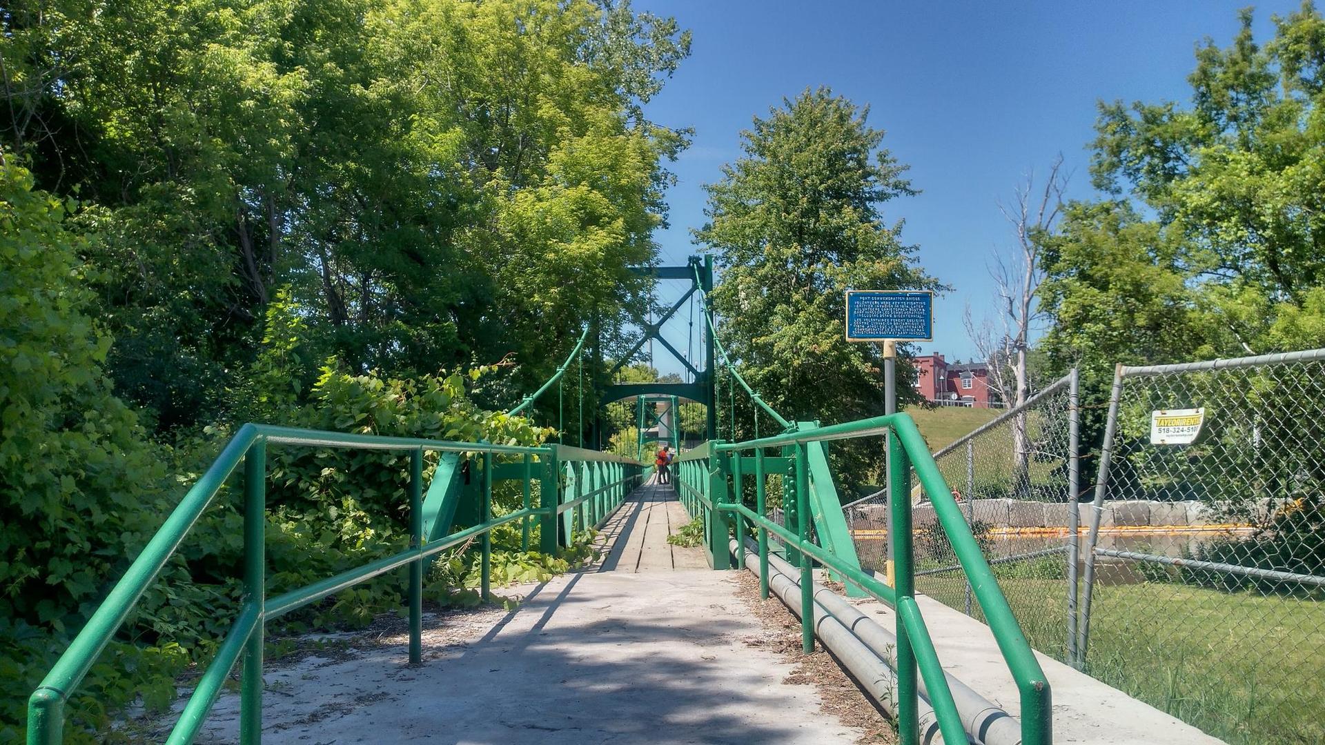 Green suspension bridge over water, surrounded by lush summer trees.