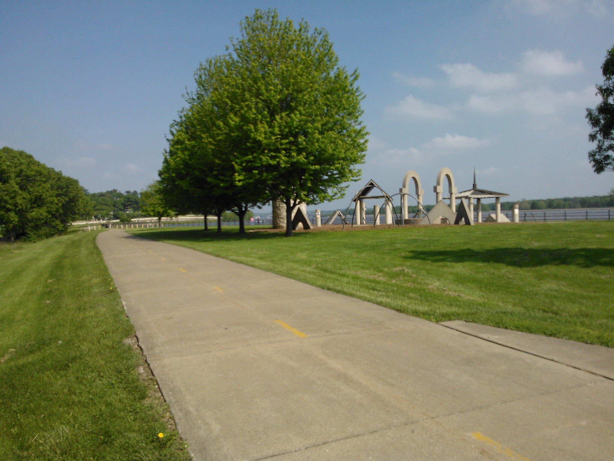 Paved path curving through a sunny green park with trees and structures. - Sylvan Island
