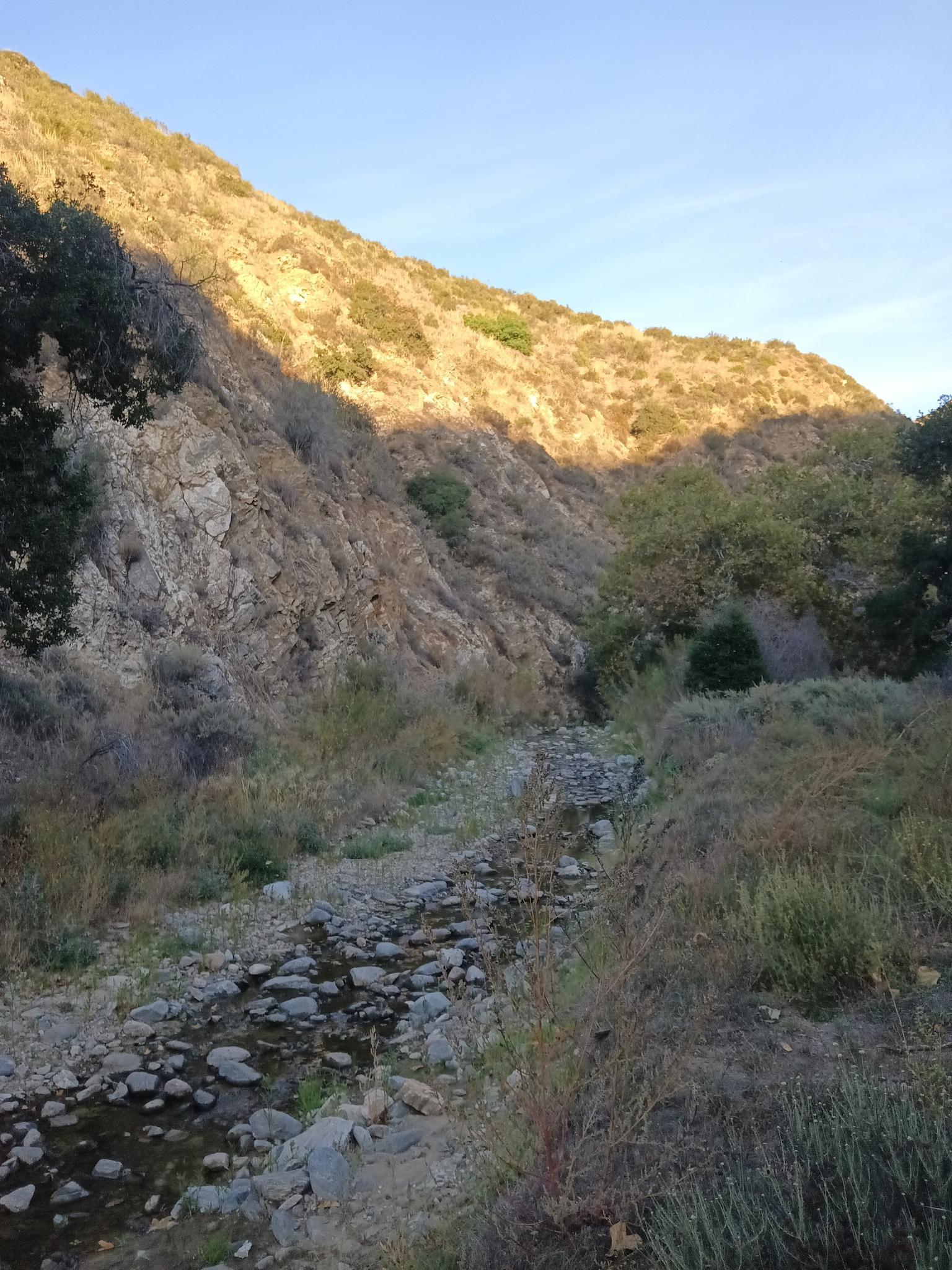 Rocky stream bed in a canyon, with sunlit hills and clear sky.