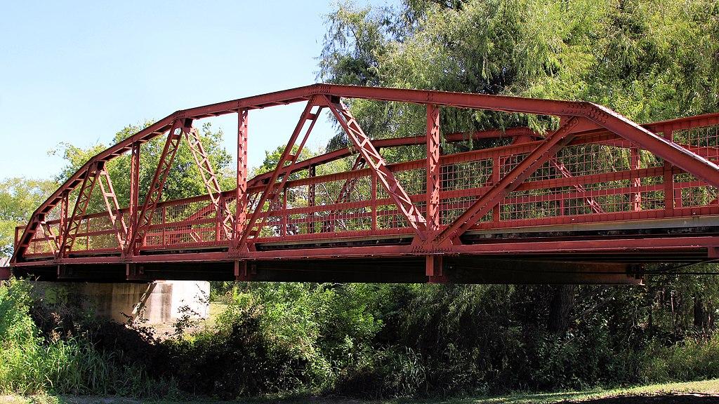 Bright red metal truss bridge spanning over green foliage on a sunny day.
