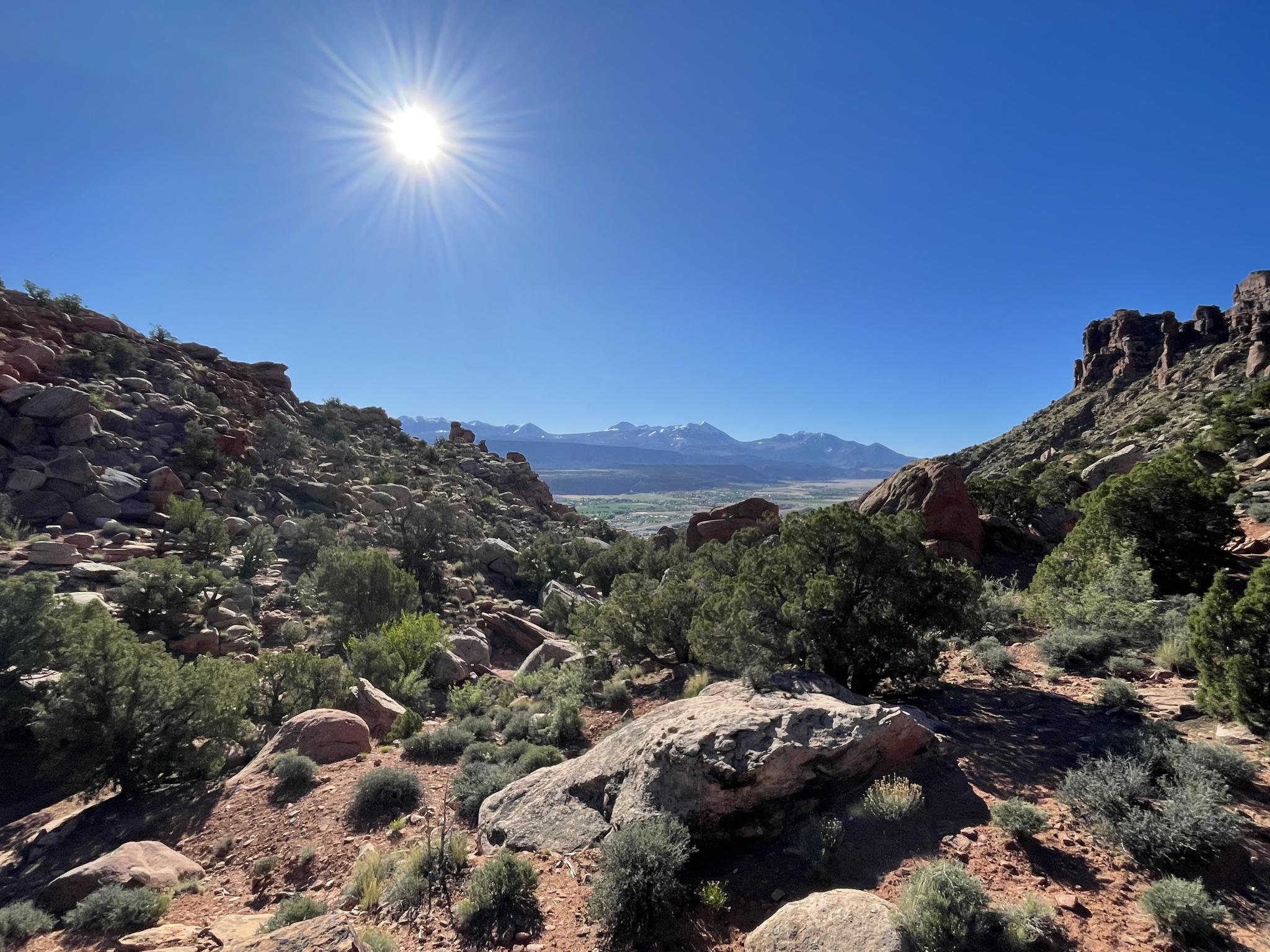 Bright sun over rocky desert landscape with distant snow-capped mountains.