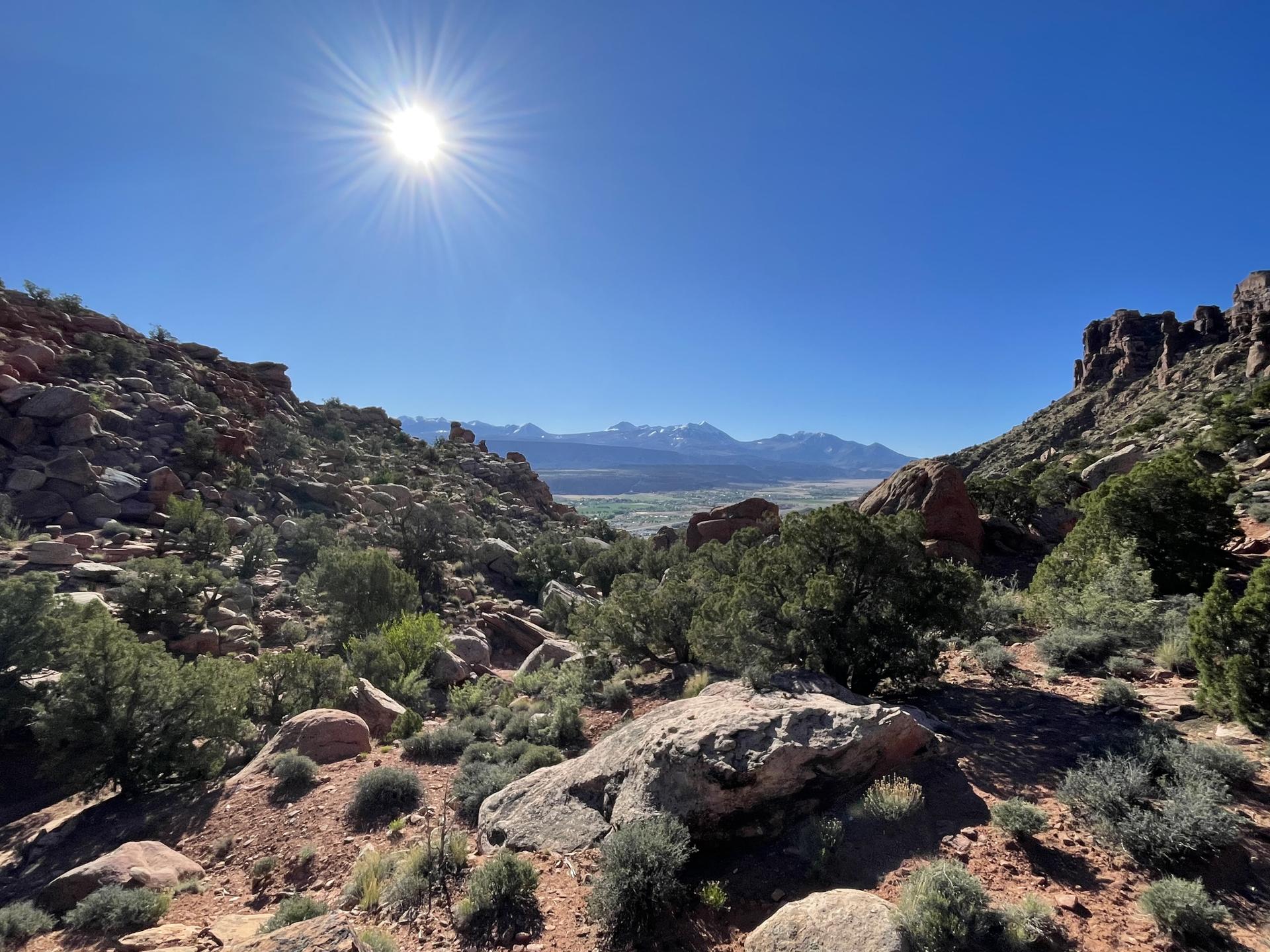 Bright sun over rocky desert landscape with distant snow-capped mountains.