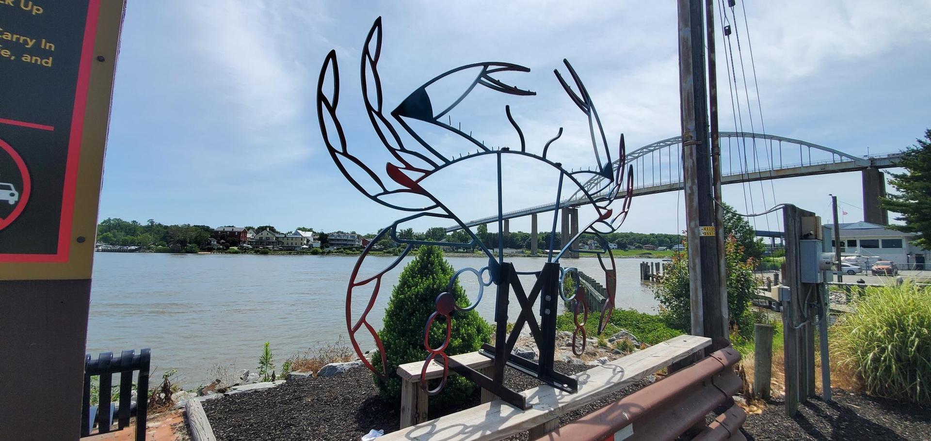 A metal crab sculpture sits by a river with a bridge in the background.