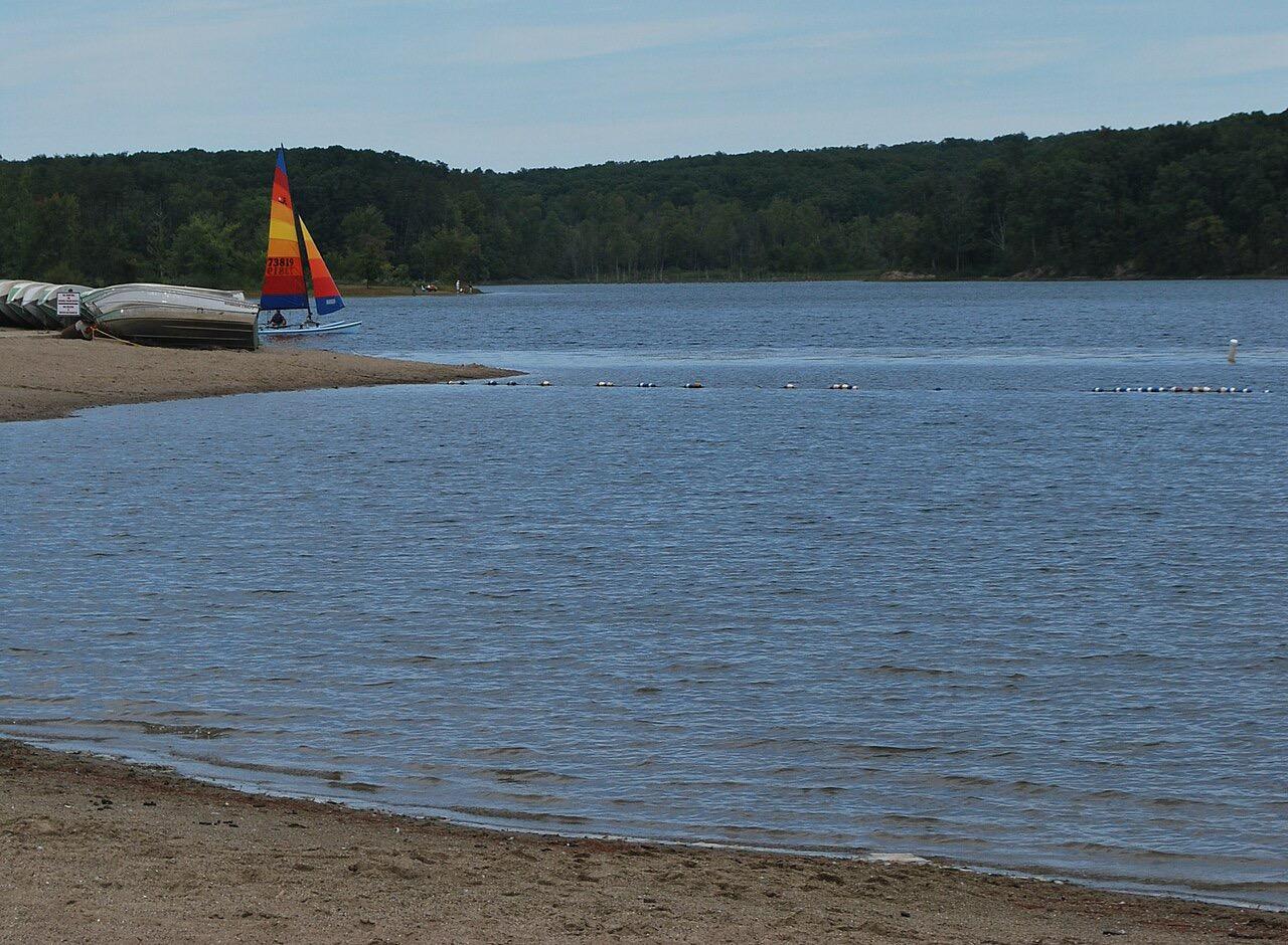 A sailboat with orange and yellow sails on a lake near a sandy beach.