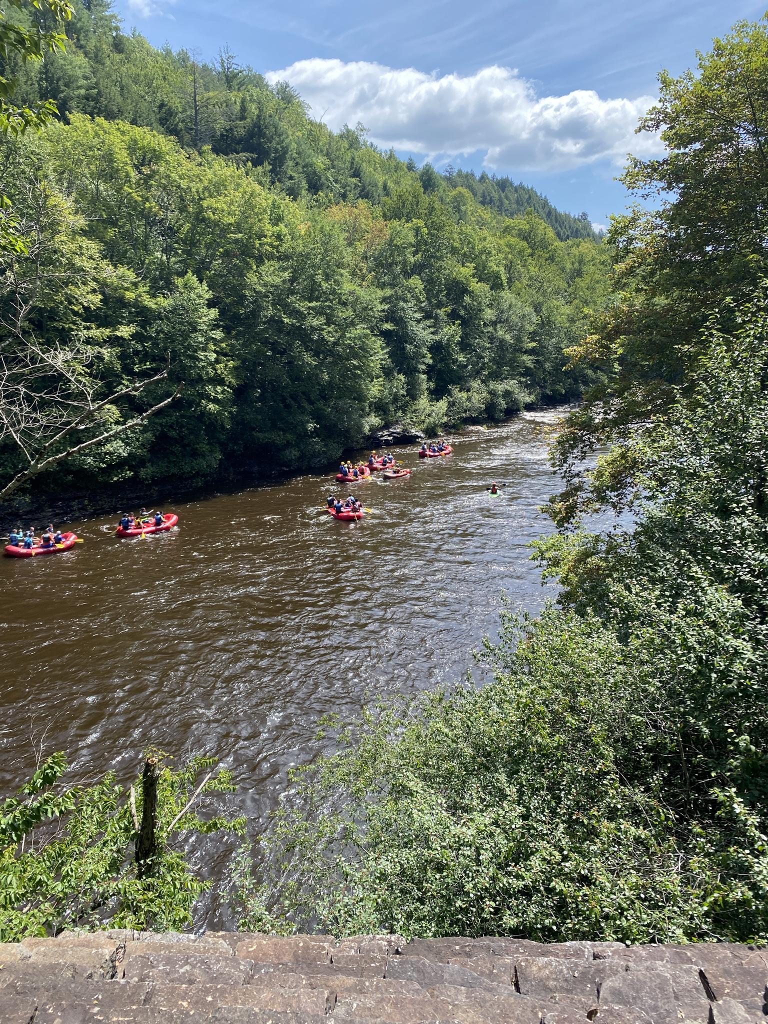 Several red rafts with people paddle down a wide river surrounded by forested hills.