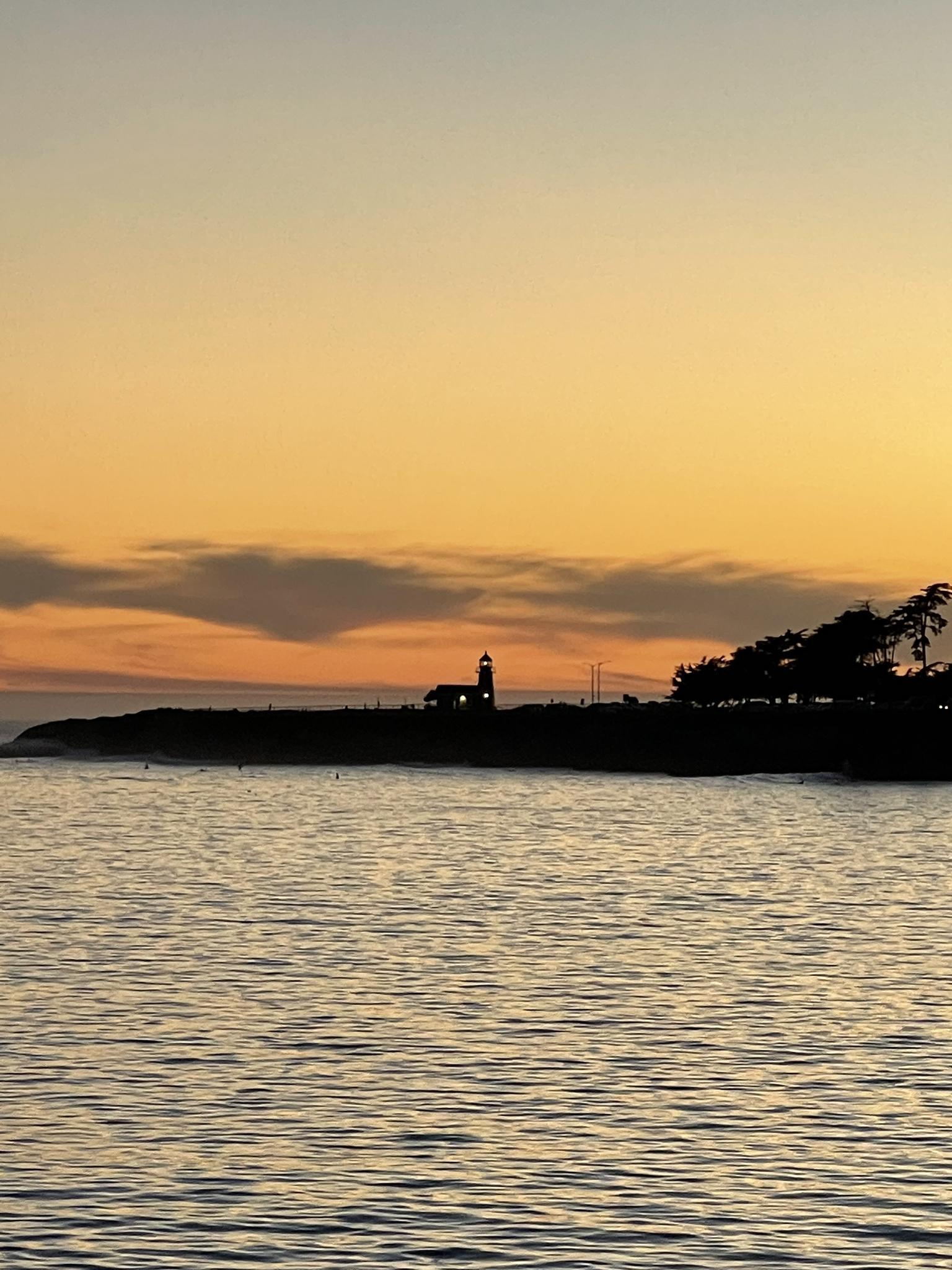 Silhouetted lighthouse and coastline against a warm orange sunset over water.