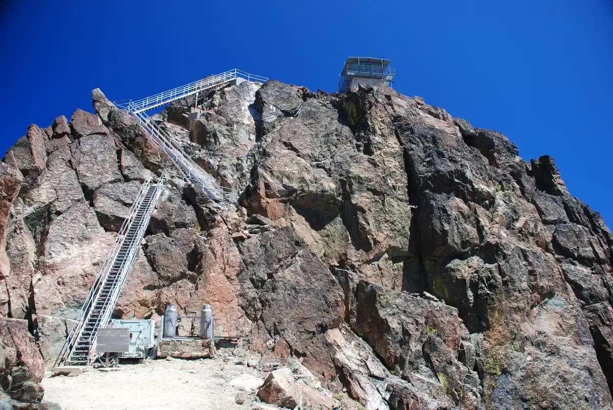 Steep rocky mountain peak with a fire lookout tower and stairs under clear blue sky.