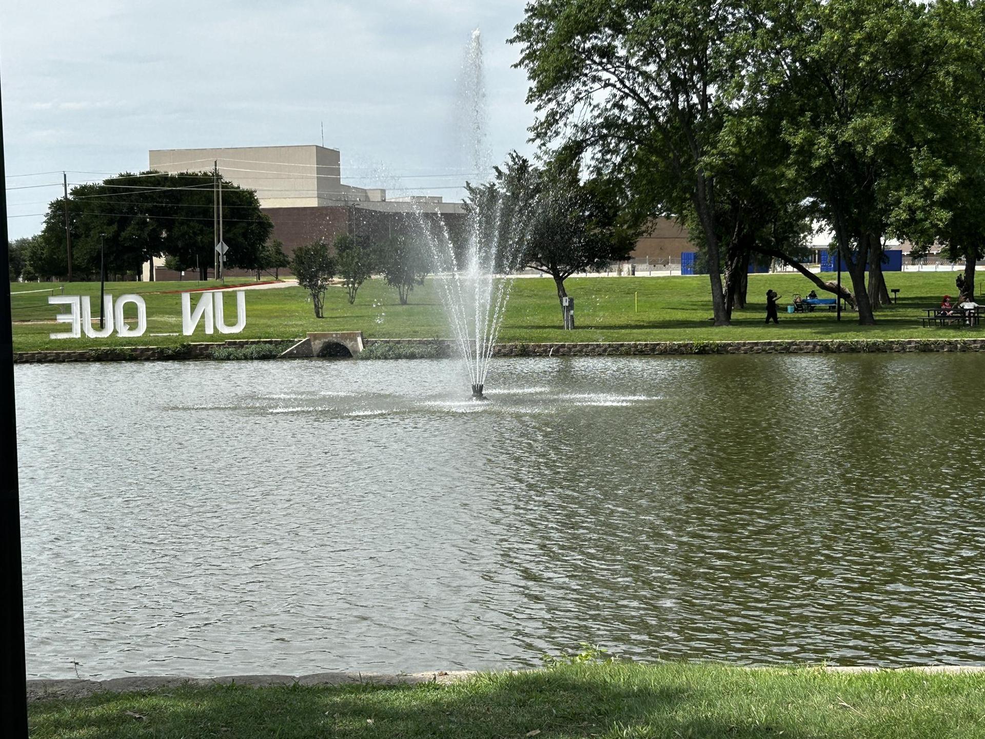 Lake with fountain, grassy banks, distant buildings, people relaxing.