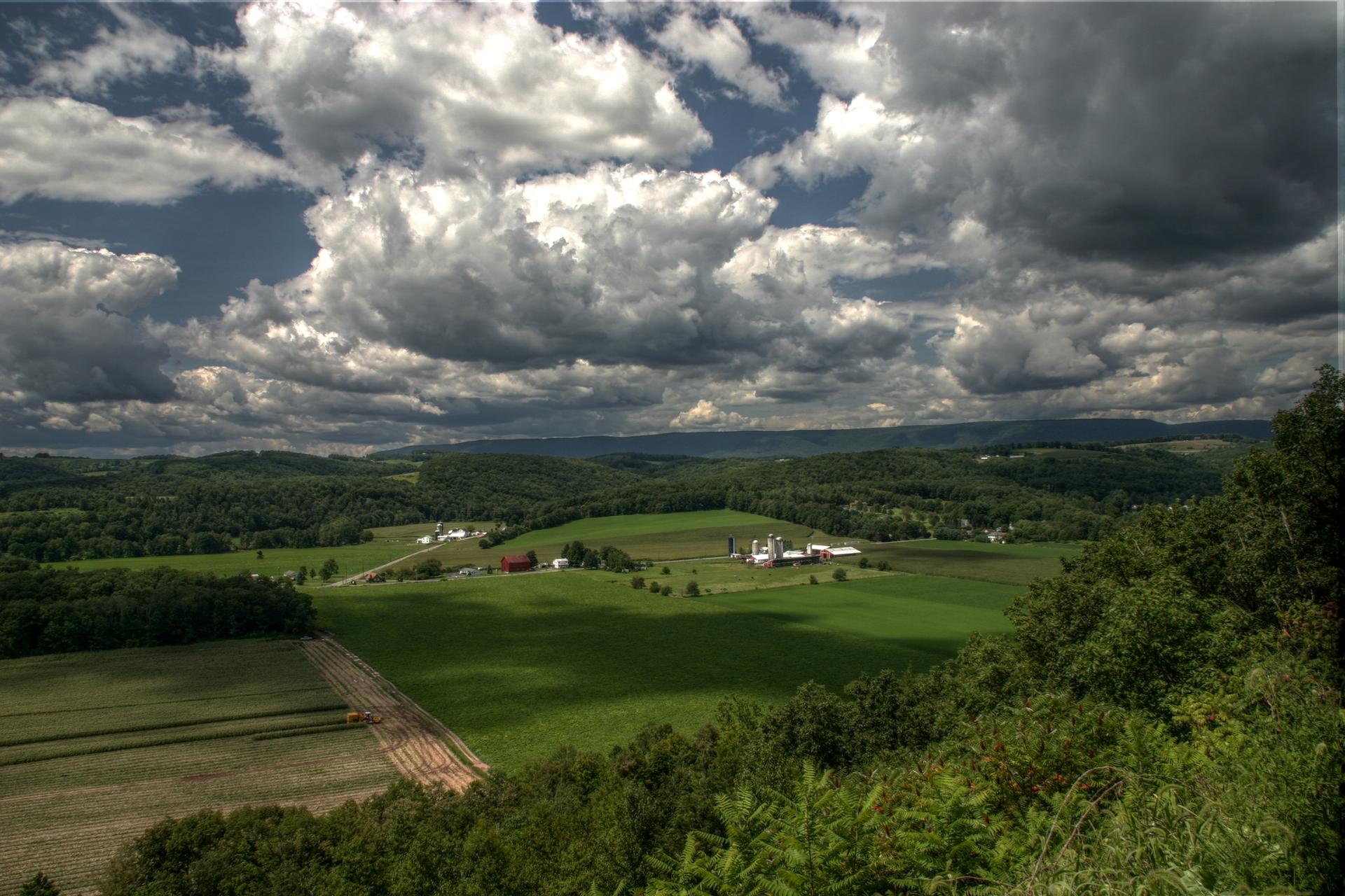 Expansive green fields and forests under a dramatic cloudy sky.