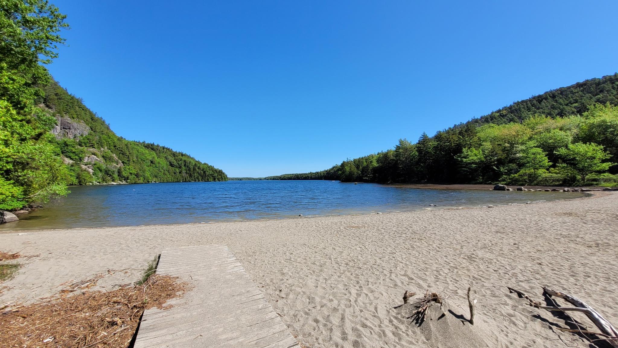 Sandy beach leading to a calm lake surrounded by green forested hills. Bright sunny day.