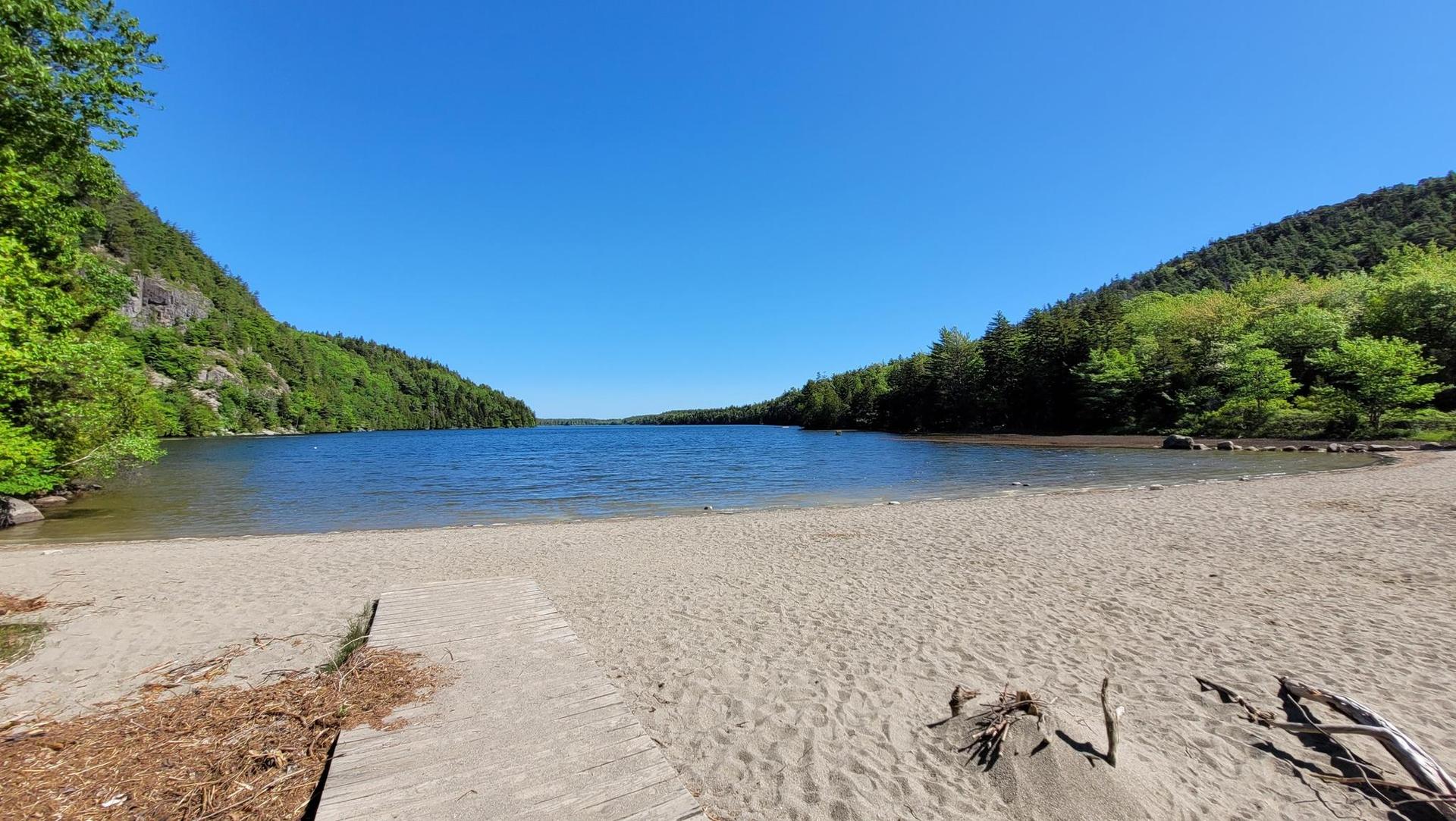 Sandy beach leading to a calm lake surrounded by green forested hills. Bright sunny day.