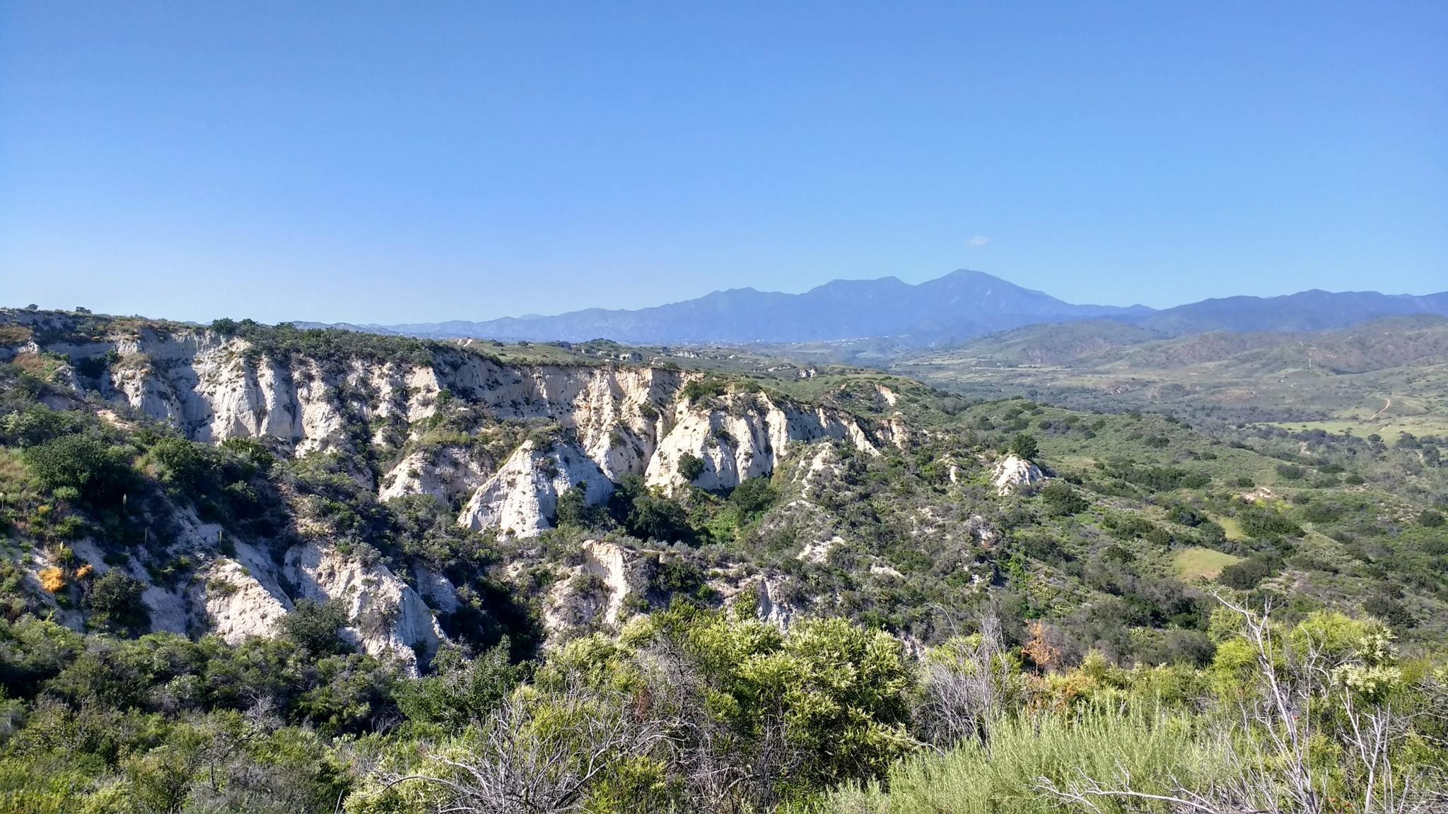 Sunny landscape with white rock formations, green hills, and distant mountains under blue sky.