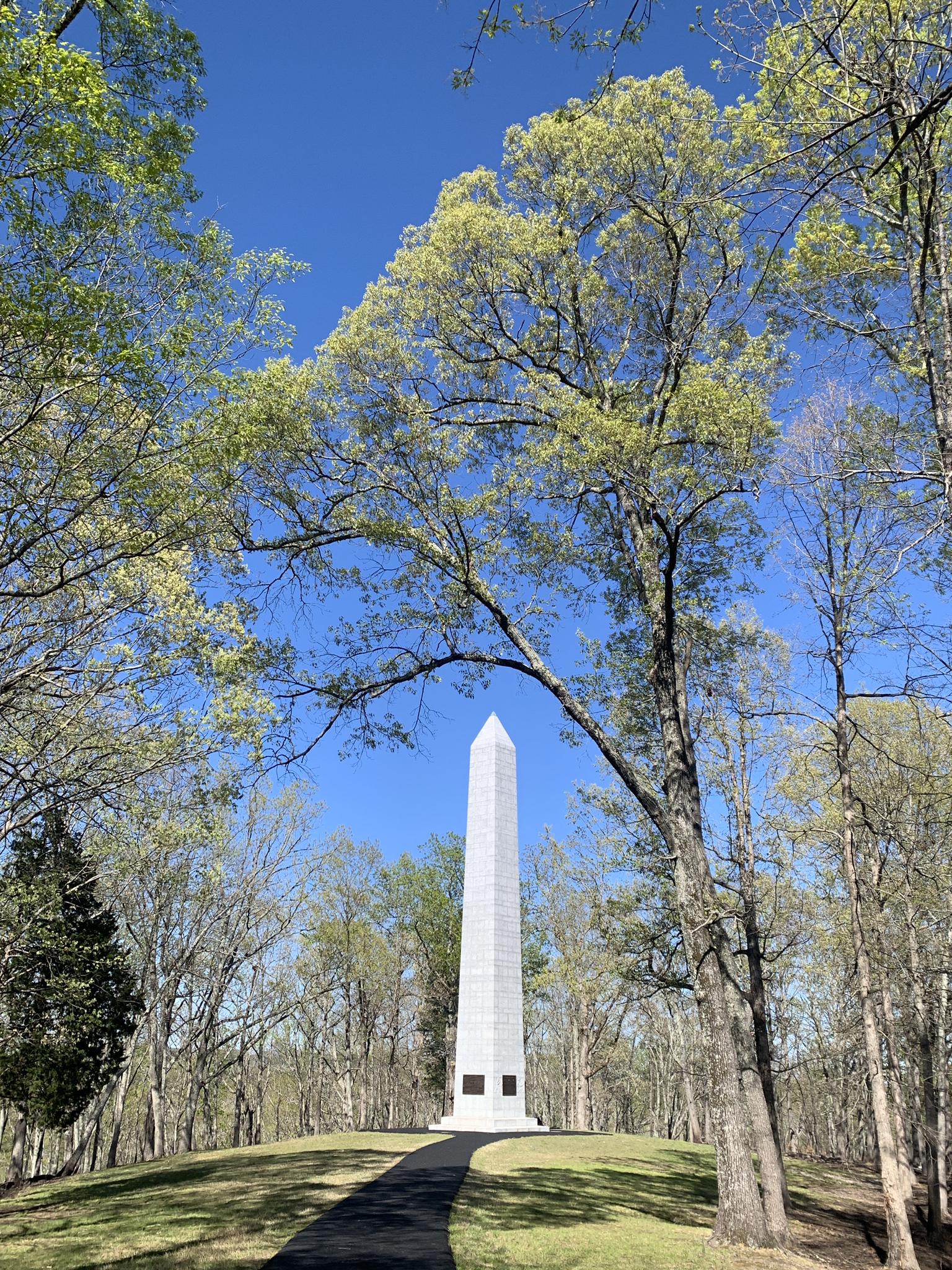 Tall white obelisk monument framed by green trees under a bright blue sky.