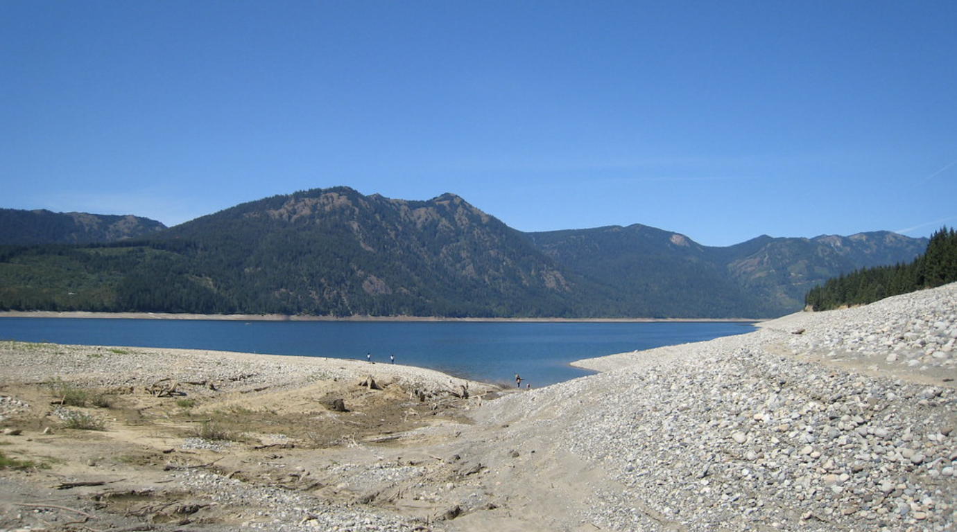 Bright lake with forested mountains, rocky shore, and distant people.